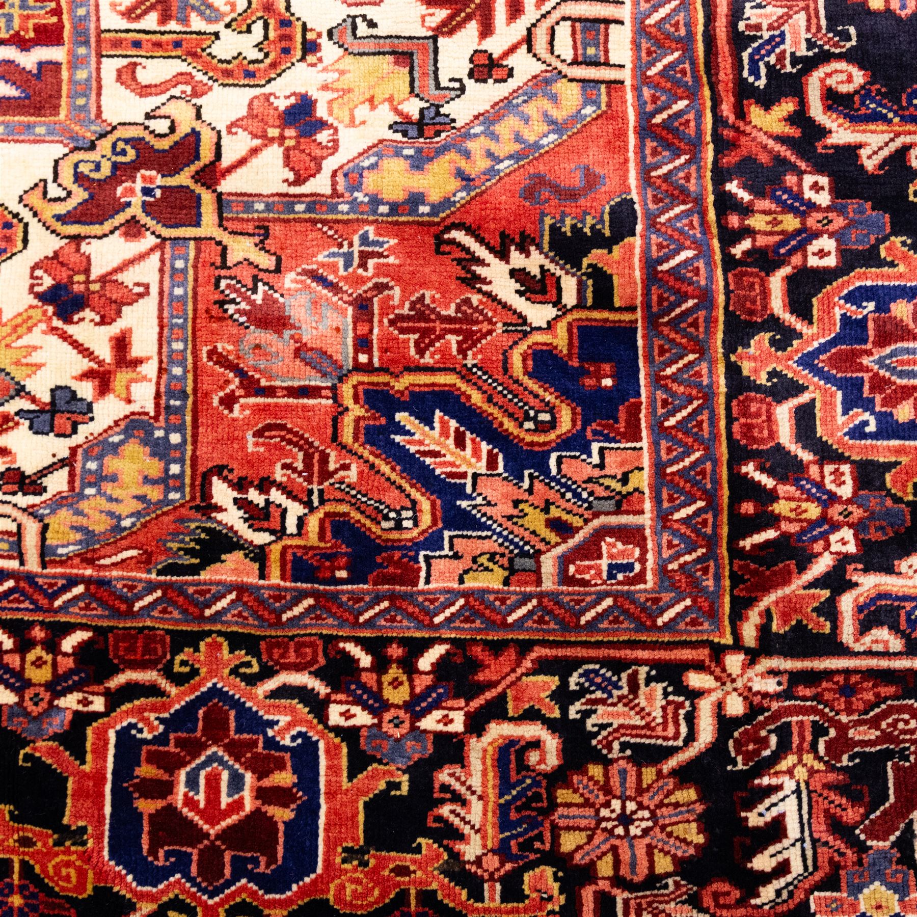 North West Persian Heriz hand-knotted carpet, madder red field with large central stepped medallion with angular floral and geometric designs, surrounded by stylized palmettes, leaves and tribal motifs with indigo corner spandrels, enclosed by a broad navy border of geometric floral devices between multiple guard stripes, with cotton fringe ends 