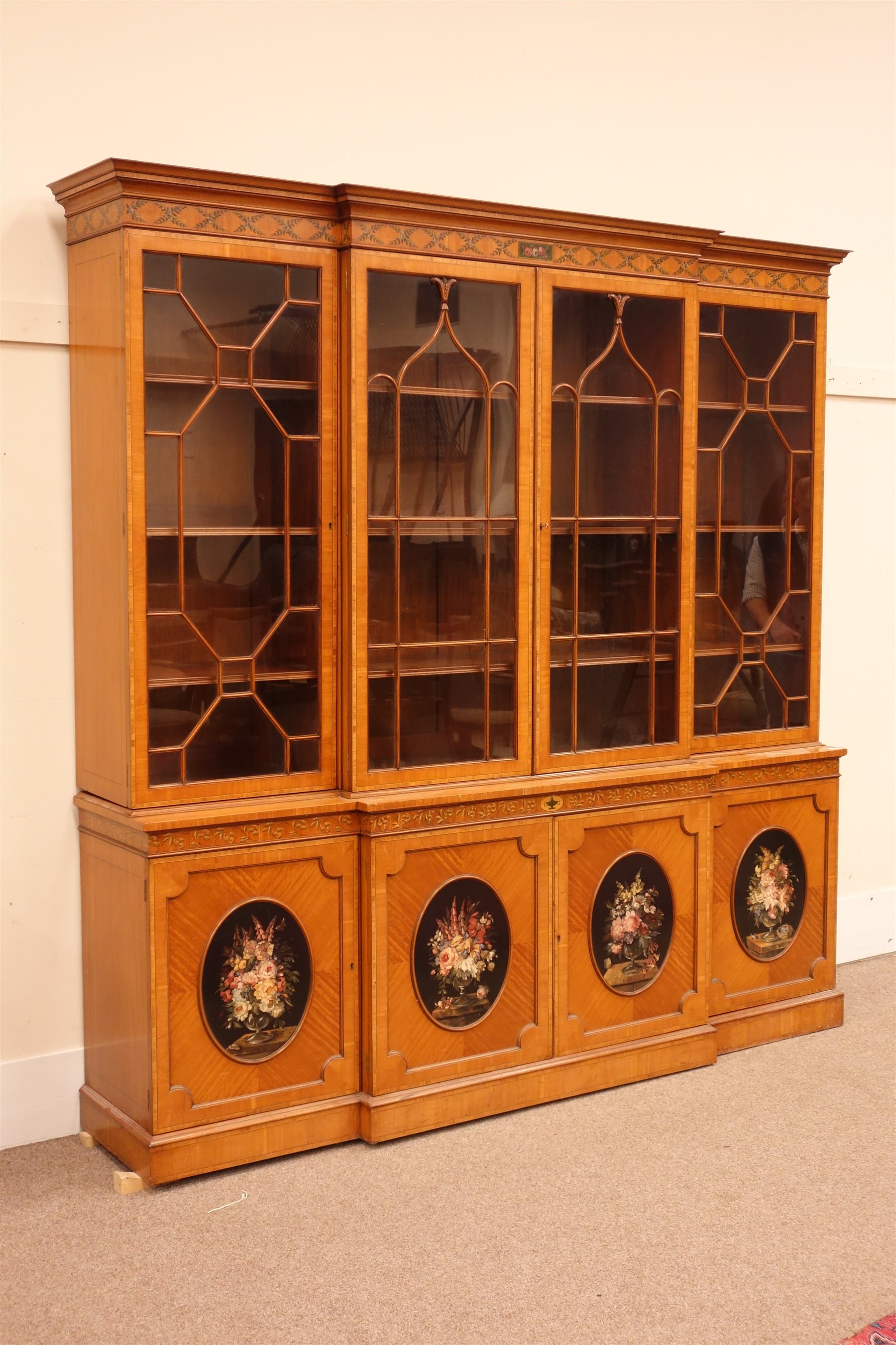 Sheraton Revival satinwood breakfront bookcase, frieze painted with scrolling leafage above four astragal doors with adjustable shelves, the base with four doors painted with floral sprays in oval panels, on a plinth base, W234cm, H235cm, D52cm