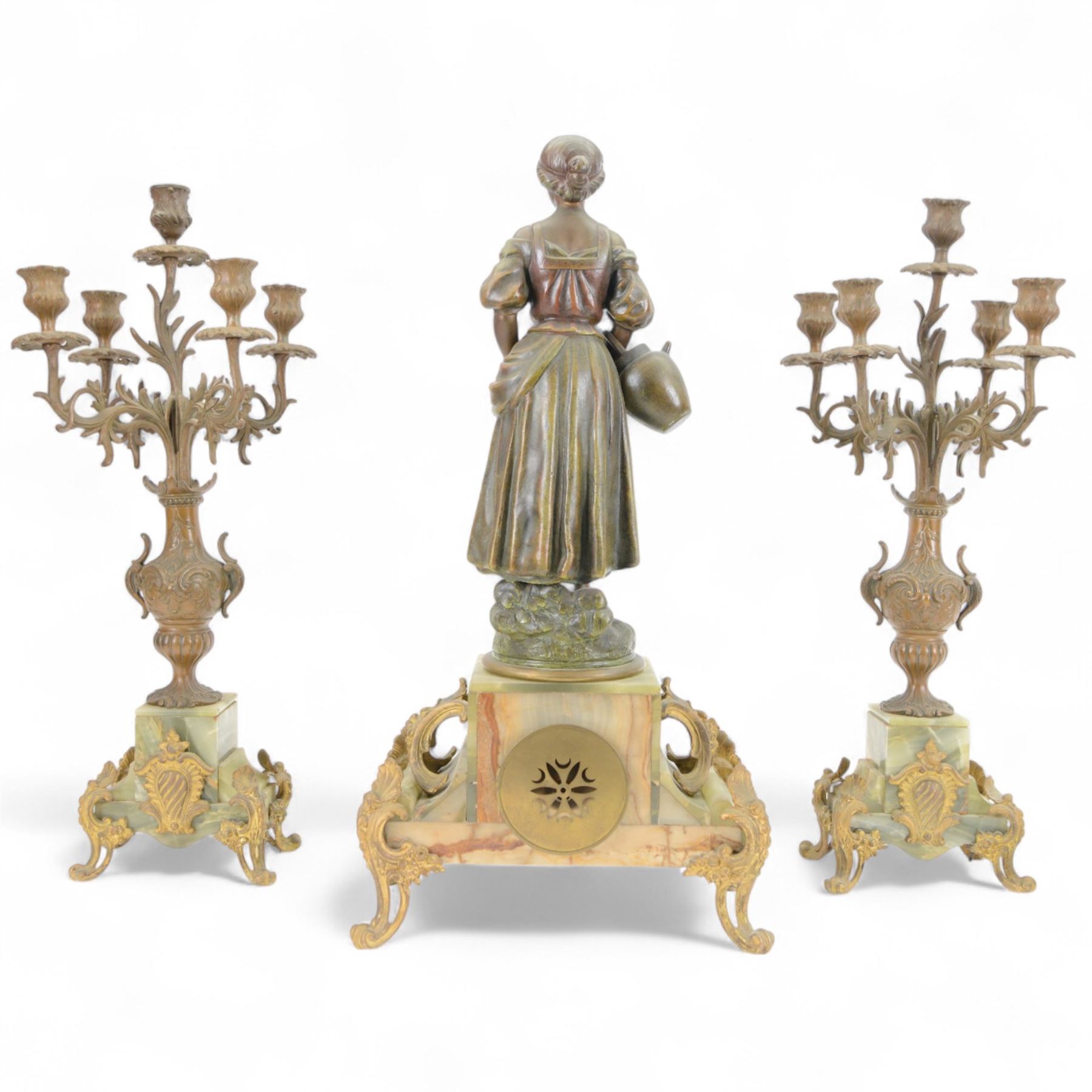 French-Edwardian green onyx clock garniture with a shaped case surmounted by a cast patinated figure of  a maiden in 18th century dress, with gilded scroll feet and conforming mounts, porcelain dial with Arabic numerals and floral decoration, twin train rack striking movement sounding the hours and half-hours on a bell, with a matching pair of five light candelabra. With pendulum.    
