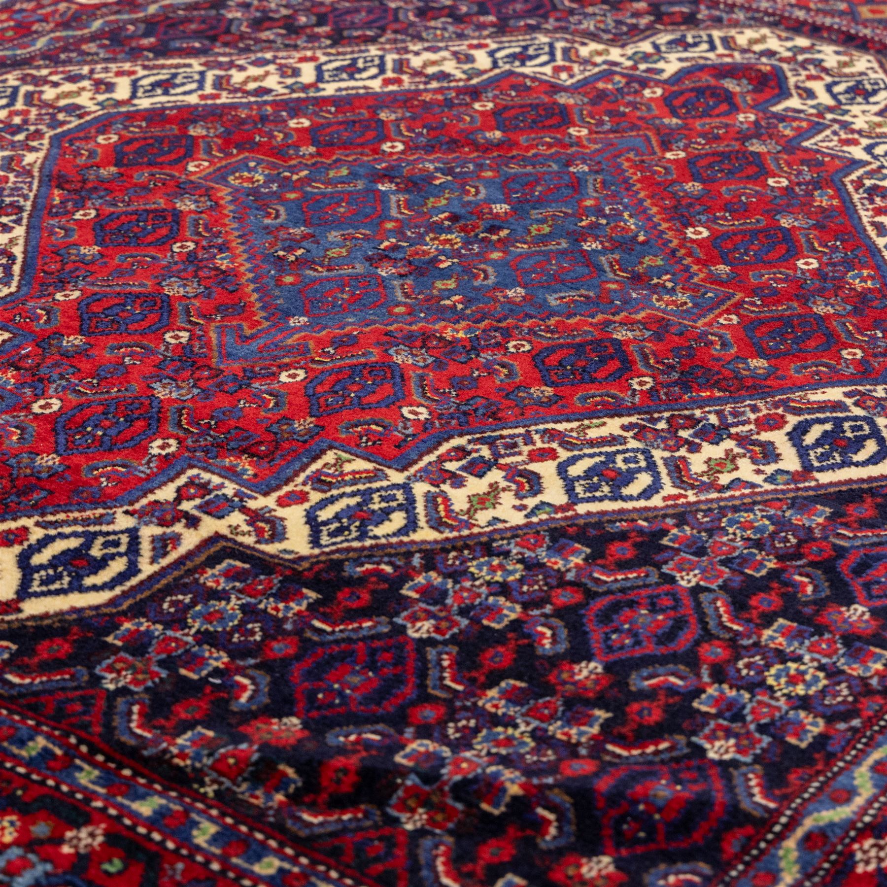 North West Persian Senneh hand-knotted rug, dark indigo field with a central bold stepped lozenge medallion of madder red and deep blue, enclosed within an ivory serrated contour forming a cruciform outline, medallion with repeating boteh and floral devices, field decorated with an all-over Herati pattern of rosettes enclosed by angular leaves principal border of madder red ground decorated with alternating floral heads, flanked by narrow guard stripes, with cotton fringe ends 