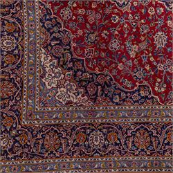 Central Persian Kashan hand-knotted carpet, madder red field with central cusped medallion in navy and ivory enclosing a floral rosette and radiating scrolling arabesques and palmettes, across a densely ornamented ground of finely drawn vinery enriched with a profusion of flowering sprays and stylized blossoms, spandrels in deep indigo and principal border of navy ground decorated with a continuous palmette and vine-scroll flanked by multiple guard stripes, with cotton fringe ends