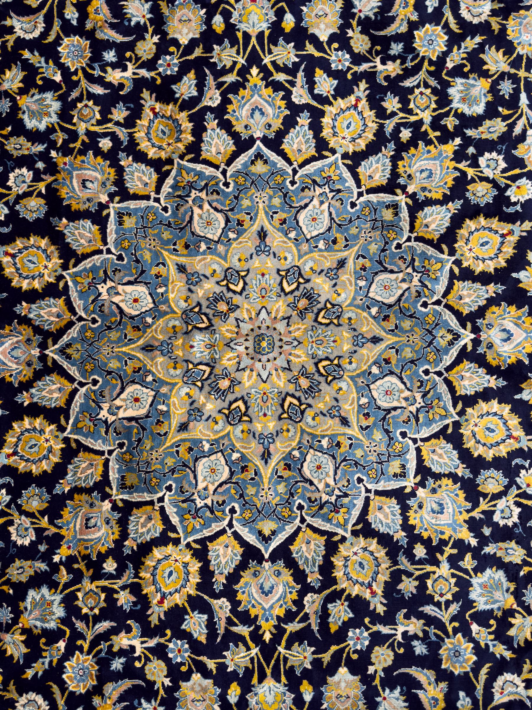 Central Persian Kashan hand-knotted carpet, the navy field with central medallion in pale blue and gold tones, surrounded by spiralling arabesques and palmettes, framed by quarter spandrels and a wide border of stylised lotus and rosette motifs between floral guard stripes, from the region of Kashan