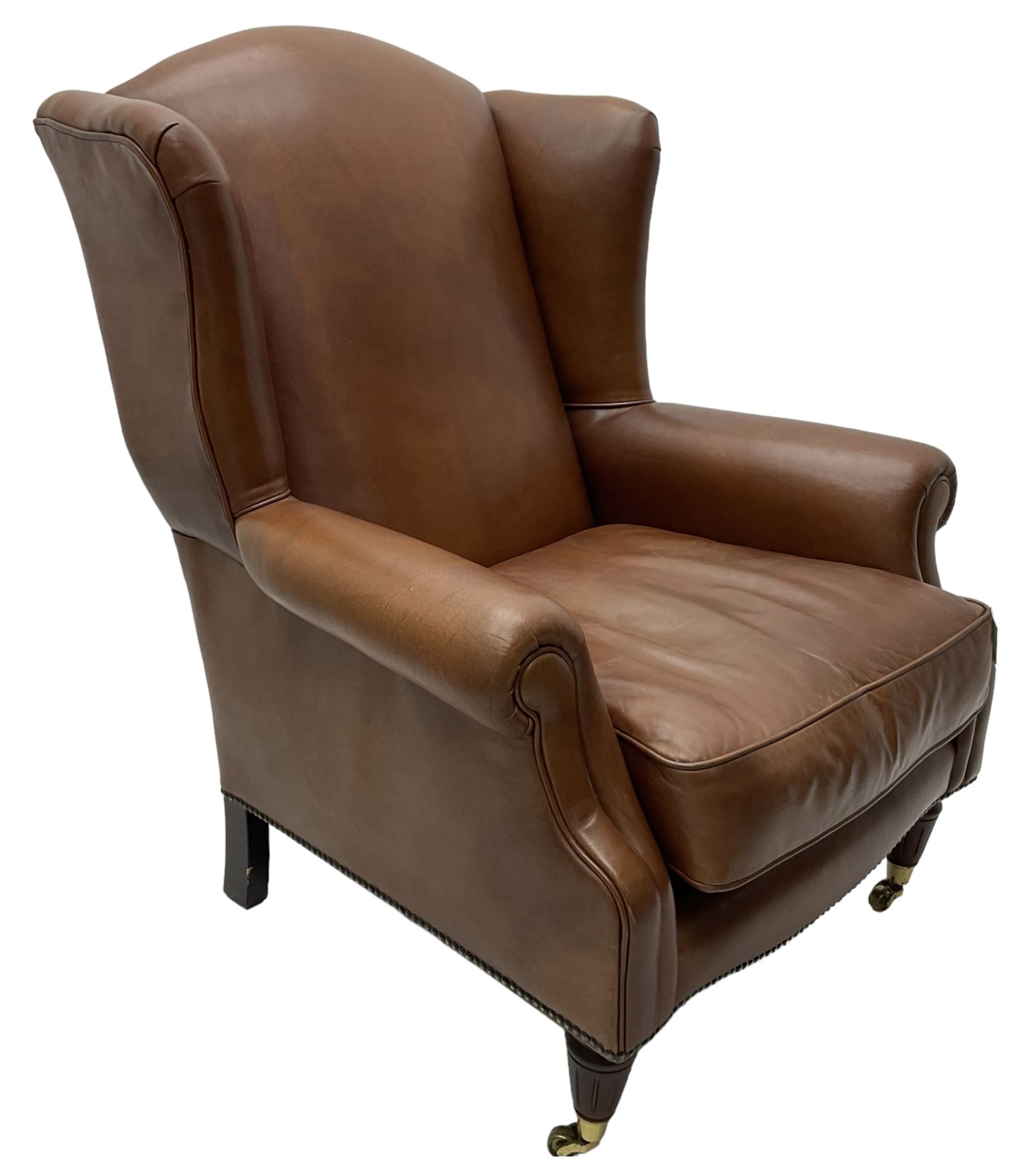 Georgian design wingback armchair, upholstered in brown leather with stud band and piping, turned and fluted front feet on brass cups and castors