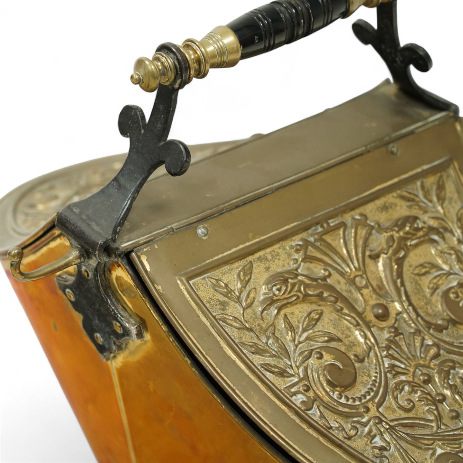Late 19th century brass coal scuttle, of helmet form, with embossed scrolling foliate panels, cast iron supports with turned ebonised handle, raised on a cast iron tripod base