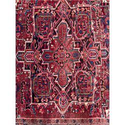 Northwestern Persian Heriz hand-knotted carpet, red madder field with a bold stepped medallion in indigo, ivory, and terracotta, flanked by angular floral motifs and stylized leaves, the spandrels set within a dark indigo border of rosettes and angular vine motifs between narrow guard stripes, 340cm x 245cm 