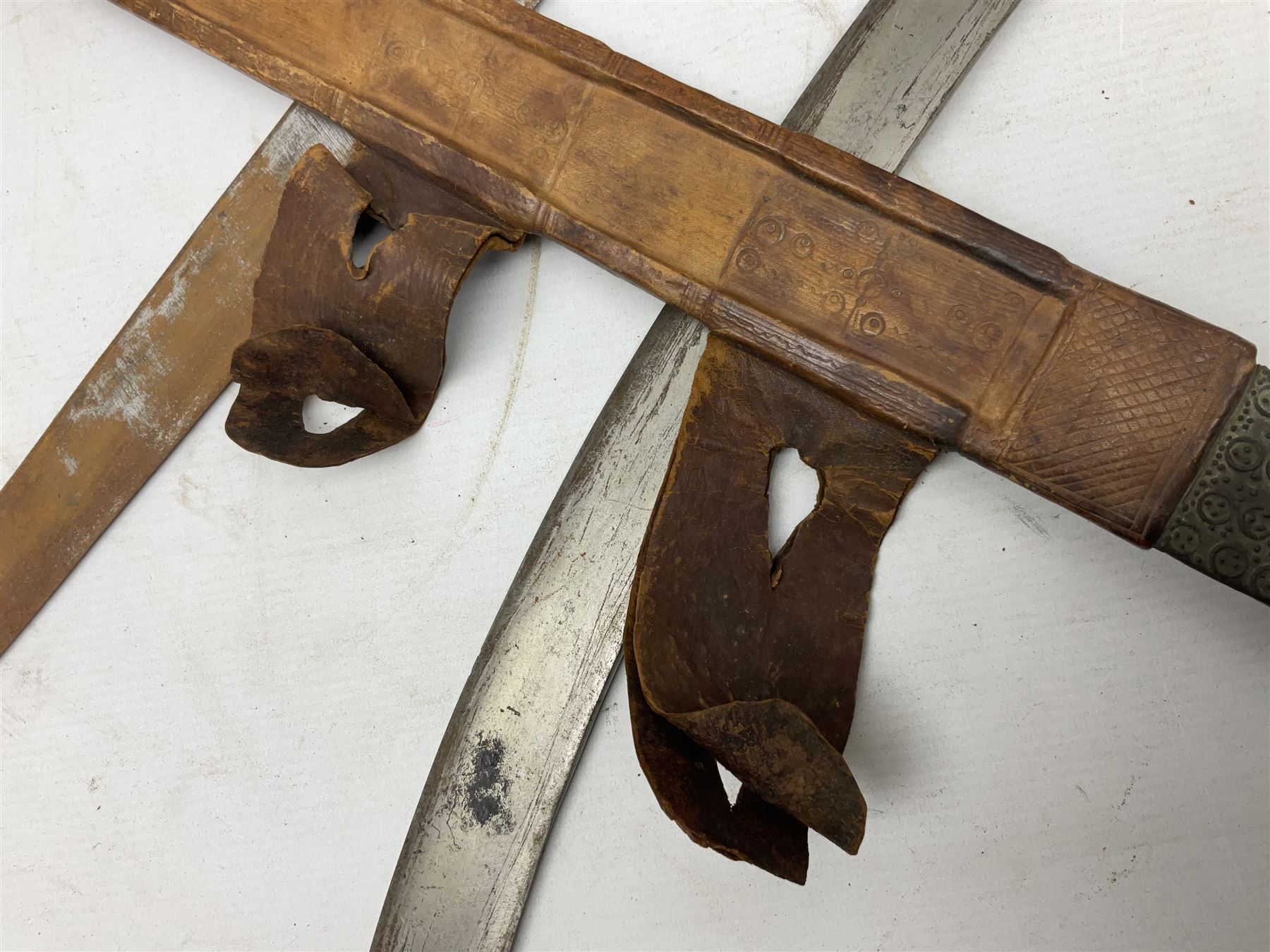 Tuareg Takoba sword with 78.5cm triple fullered blade incised with two crescent moons to either side, metal hilt with stylised floral designs to the cross-guard, decorative grip and disc form pommel cap; in tooled leather scabbard decorated with geometric designs L92cm overall; and an Indian Tulwar sword with 75cm plain curving blade and 'silver' damascened hilt (no scabbard) (2)