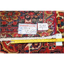 North West Persian Heriz/Ahar hand-knotted carpet, brick-red field with a large central angular stepped medallion with pendant motifs, surrounded by stylized palmettes, serrated leaves and geometric floral devices , ivory corner spandrels, enclosed by a dark indigo main border of stylized rosettes and angular vine motifs, between multiple guard stripes, with cotton fringe ends 