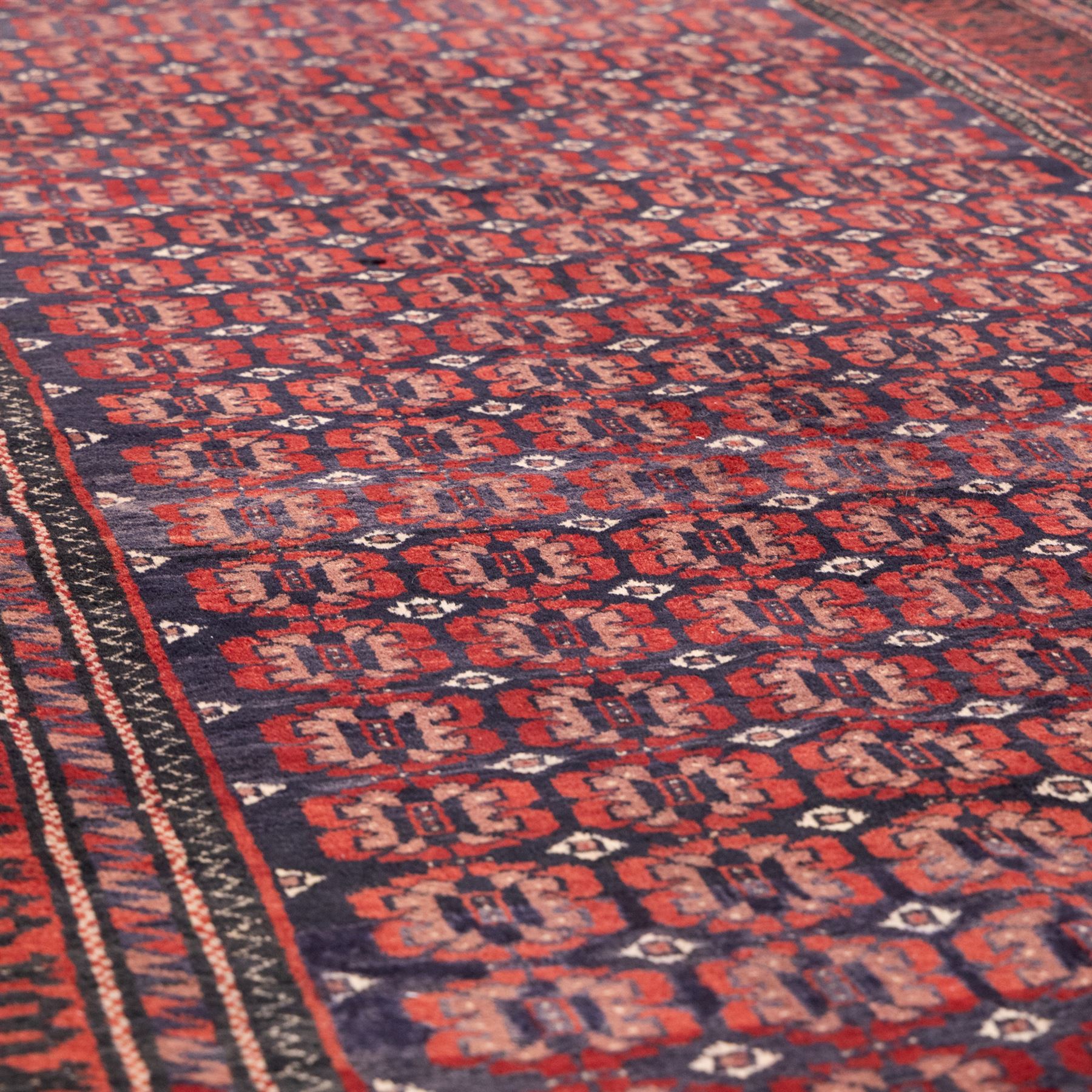 North East Persian Turkoman hand-knotted rug, indigo field densely decorated with an all-over repeating design of geometric gul motifs in shades of madder red, aubergine, and dark brown, highlighted with small ivory details, framed by multiple narrow guard borders and a main border with angular geometric motifs flanked by zig-zag borders, with cotton fringe ends 