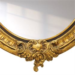 Pair of Rococo Revival gilt framed oval wall mirrors, the exuberant pediment formed of scrolling acanthus leaves and rocaille ornament, the oval bevelled plate set within with ebonised slip with outer repeating lappet decoration, over a rocaille cartouche terminal
