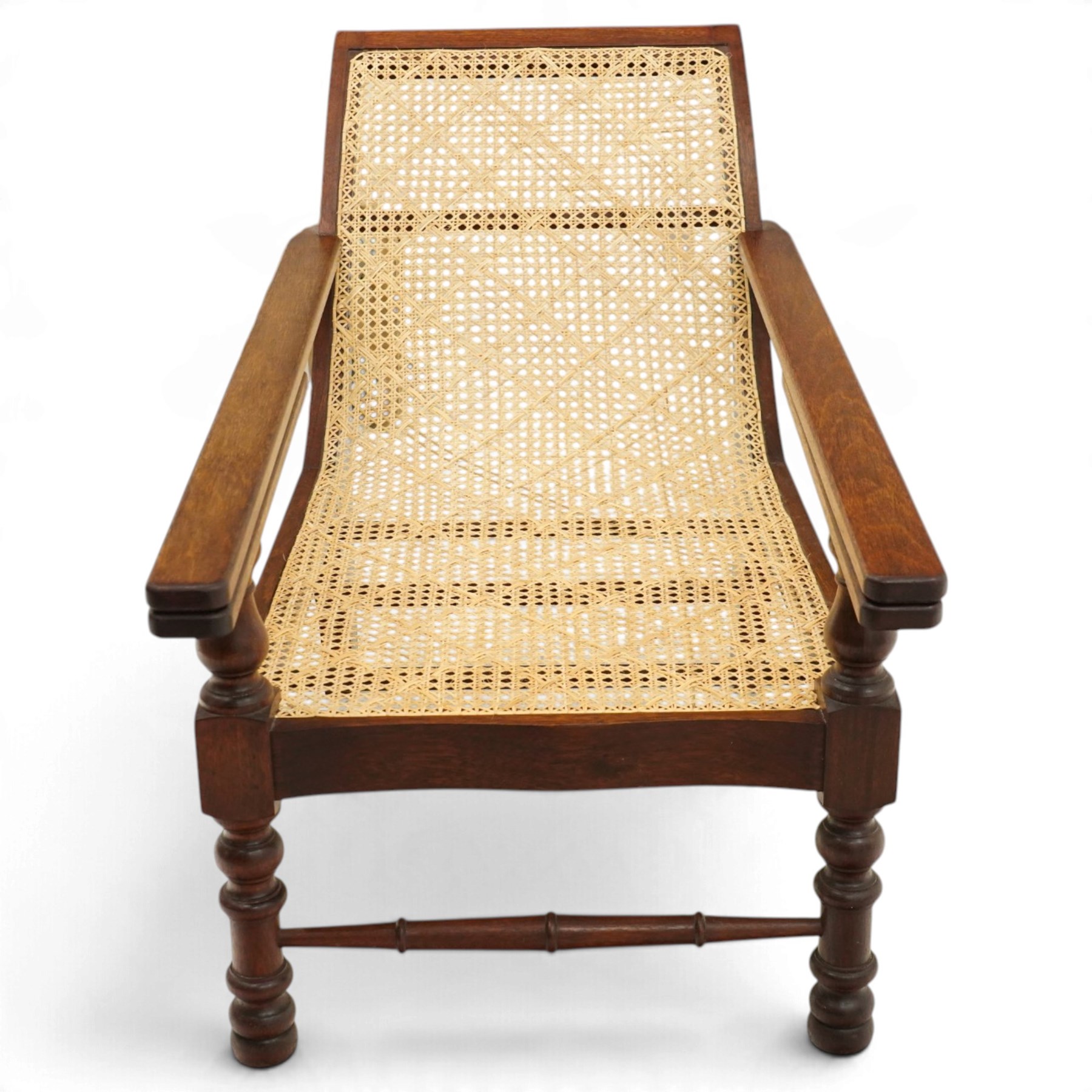 20th century teak framed plantation chair, plain cresting rail over cane back and seat, fitted with hinged extending leg rests, raised on turned front-supports united by ring-turned stretcher