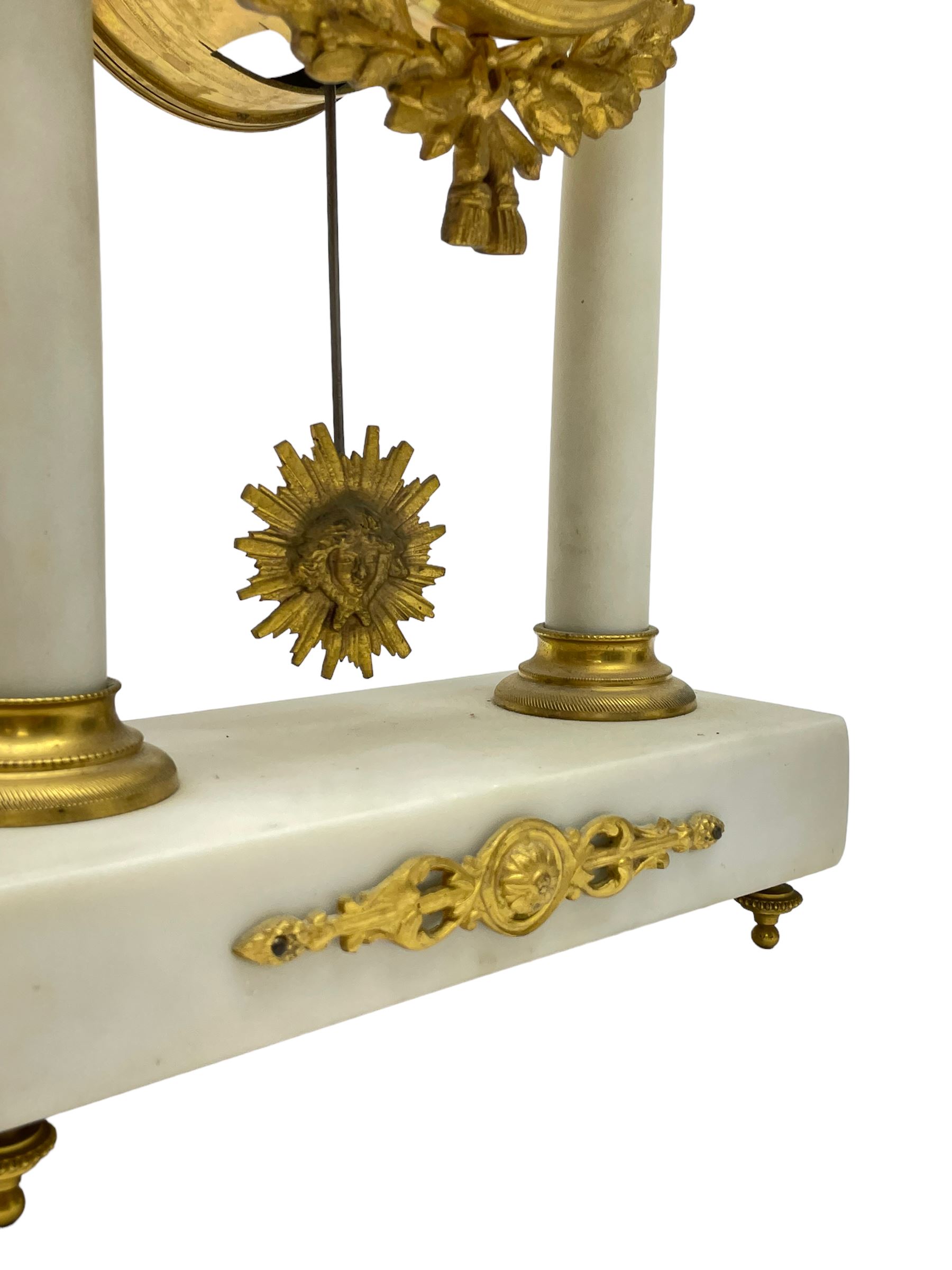 French - Late 19th century 8-day white marble and gilt metal portico clock garniture, the clock surmounted by an urn and raised on a pair of cylindrical pillars above a rectangular plinth base, white enamel dial with Arabic numerals and floral garlands, twin train countwheel striking movement, striking the hours and half hours on a bell, with a sunburst pendulum and gilt Louis XVI hands, garniture with matching twin light candelabra. With Key.   Candelabra H 30cm.