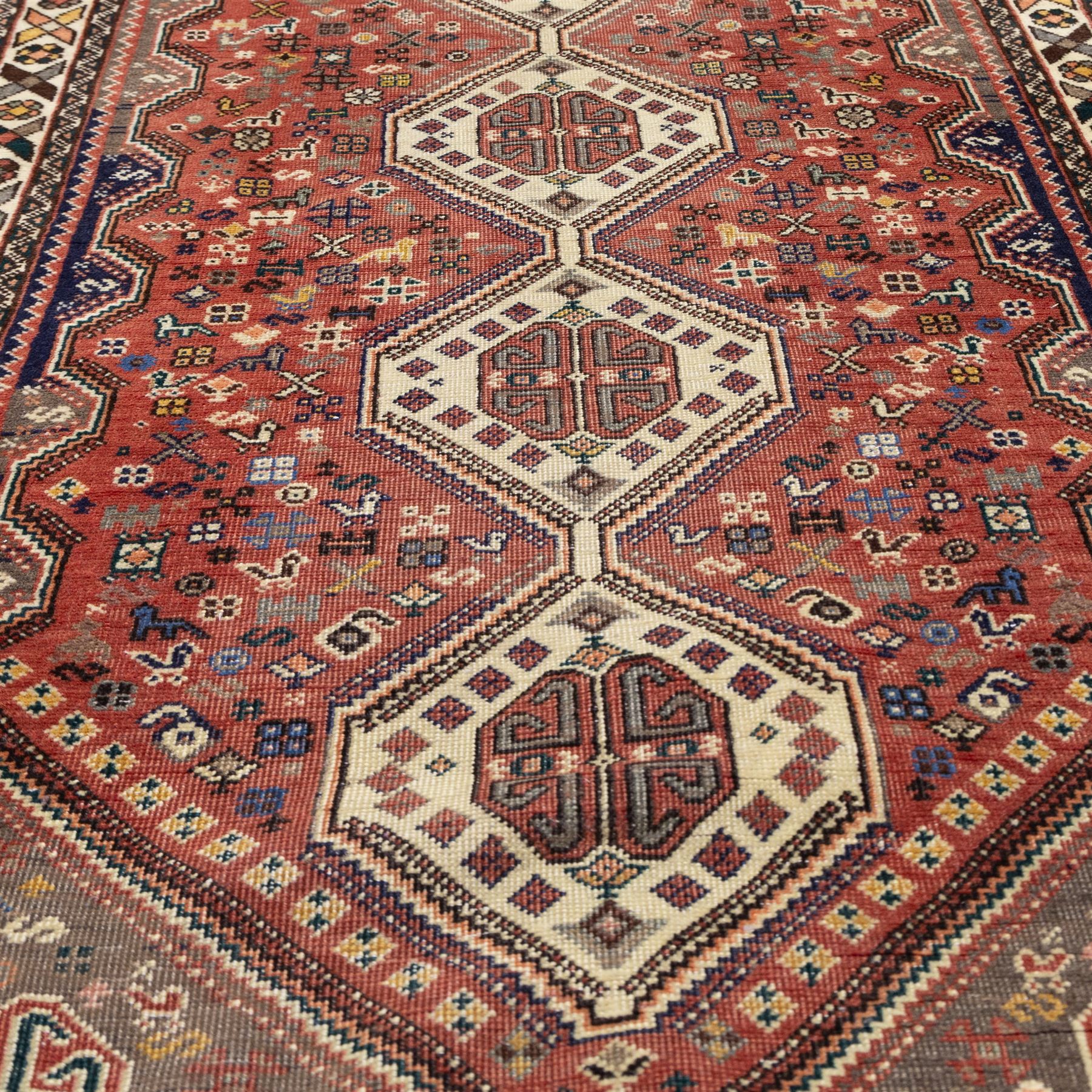 South West Persian Abadeh hand-knotted runner, madder red field decorated with a vertical sequence of stepped lozenge medallions, each enclosing stylized geometric motifs and angular devices, surrounding field filled with tribal symbols and abstract forms, enclosed within a narrow main border and flanked by multiple guard stripes, with fringe ends 