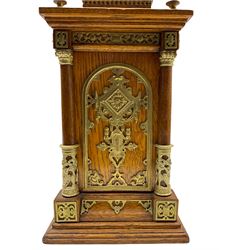 Late 19th century American mantle clock in a light oak case, raised pediment with a recessed balustrade frieze, pierced brass crown and finial, with arched rectangular conforming panels to the sides of the case, four wooden columns with capitals to each corner on a stepped plinth, circular repoussé brass dial with applied blue on white porcelain Roman numerals and Gothic steel hands, brass bezel with an egg and dart slip and flat bevelled glass, spring driven Ansonia eight-day striking movement with a recoil anchor escapement striking the hours and half-hours on a coiled gong, with fine pendulum regulation. With key and pendulum.
