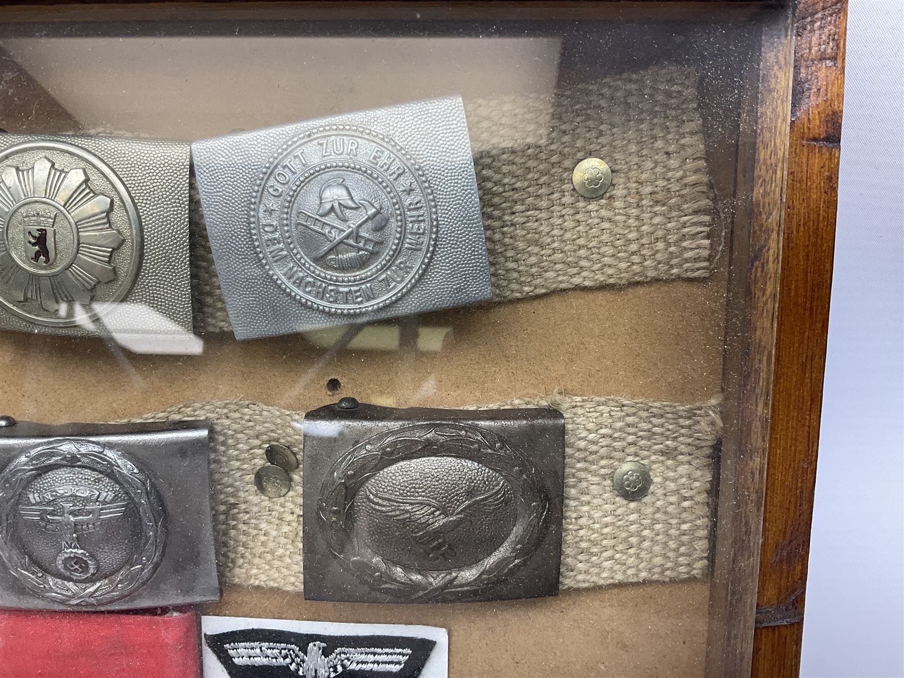 Display of eleven German belt buckles including 'SS', Party Buckle, two Army, Fire Service, two Luftwaffe, NSKK, Hitler Youth and WW1 Army; framed with three cloth badges and an armband