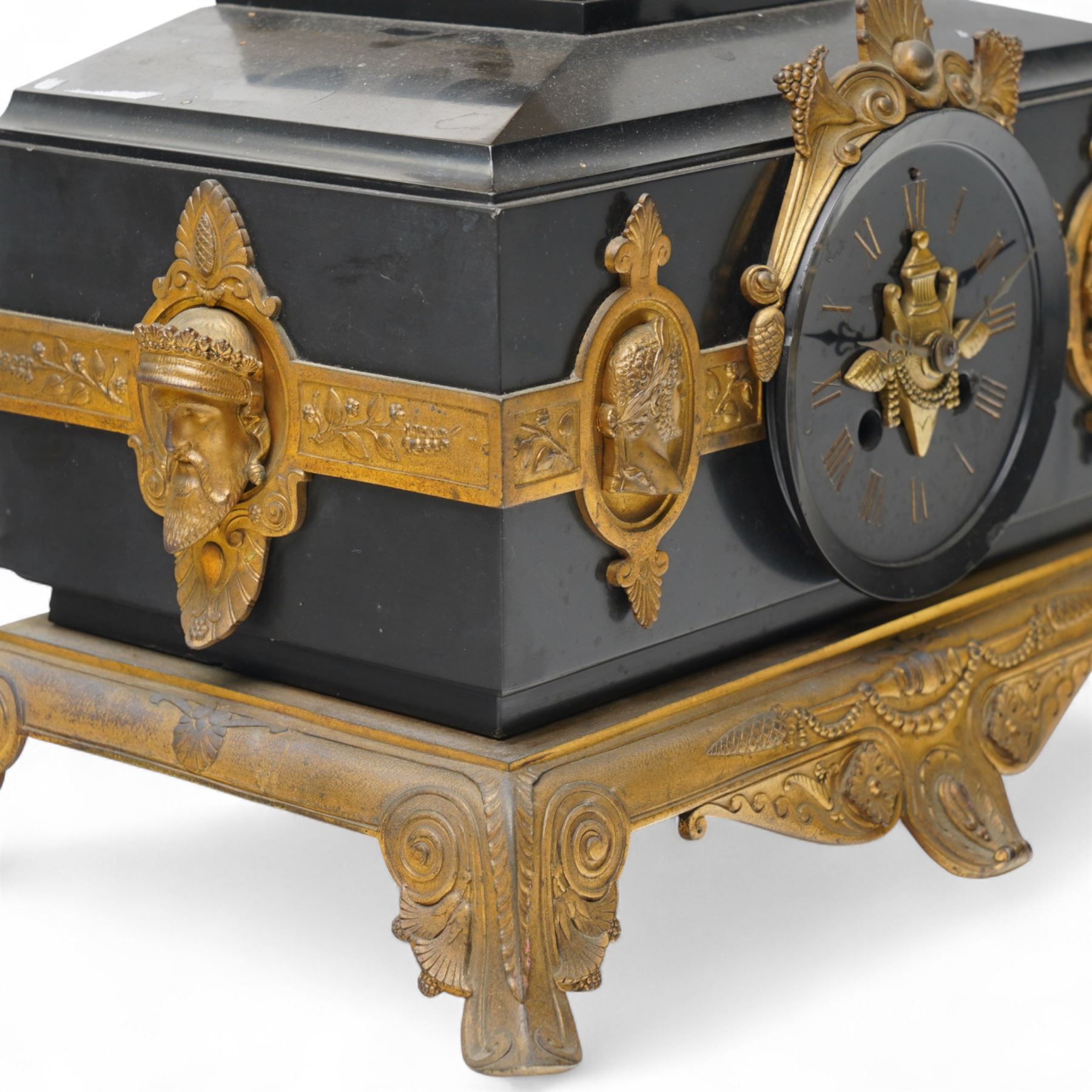French- late 19th century 8-day mantle clock in an imposing Belgium slate case with gilt metal ornamentation. Stepped rectangular case with an angled top and square plinth surmounted by an oval shaped gilt urn with scroll handles, case with continuous strapwork and oval medallions displaying portraits from classical antiquity, raised on a gilt metal base with splayed feet, conforming unenclosed slate dial with gold incised Roman numerals and brass Fleur de Lis hands, Parisian twin train rack striking movement with pendulum regulation, striking the hours and half hours on a bell. With pendulum.