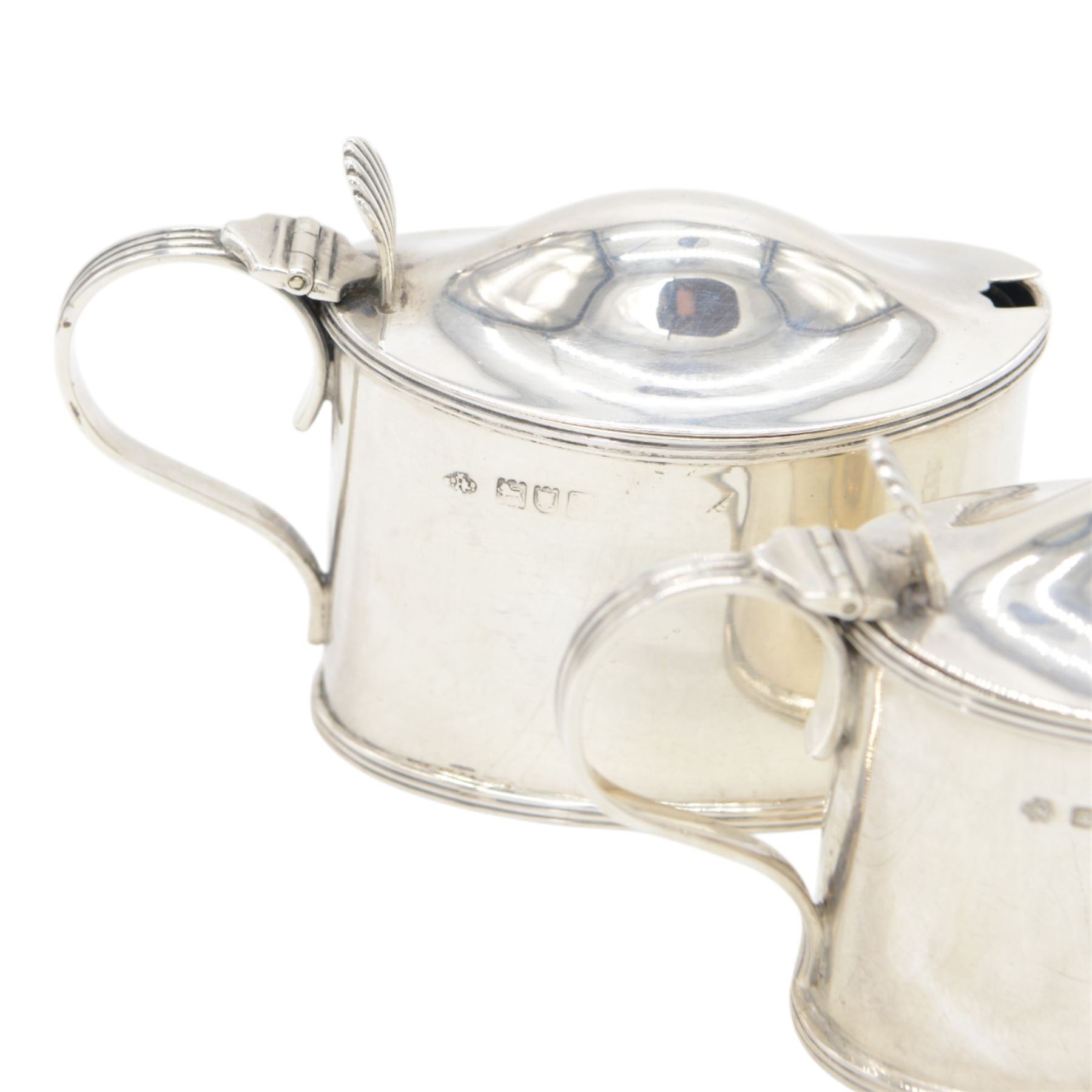 Pair of Edwardian silver mustard pots, of oval form with reed borders and palmette thumb pieces to hinged covers, opening to reveal blue glass liners, hallmarked William Hutton & Sons Ltd, London 1905, H7.5cm
