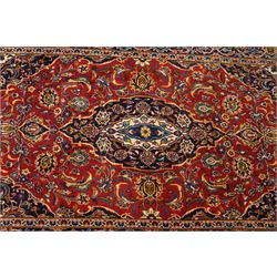 Central Persian Kashan hand-knotted rug, madder red field, with central lobed medallion with pendants, issuing an all-over design of scrolling vine, palmettes, and floral sprays, with conforming spandrels, enclosed by a broad indigo palmette and vine border framed by guard stripes in a palette of navy, blue and ivory tones, with cotton fringe ends 