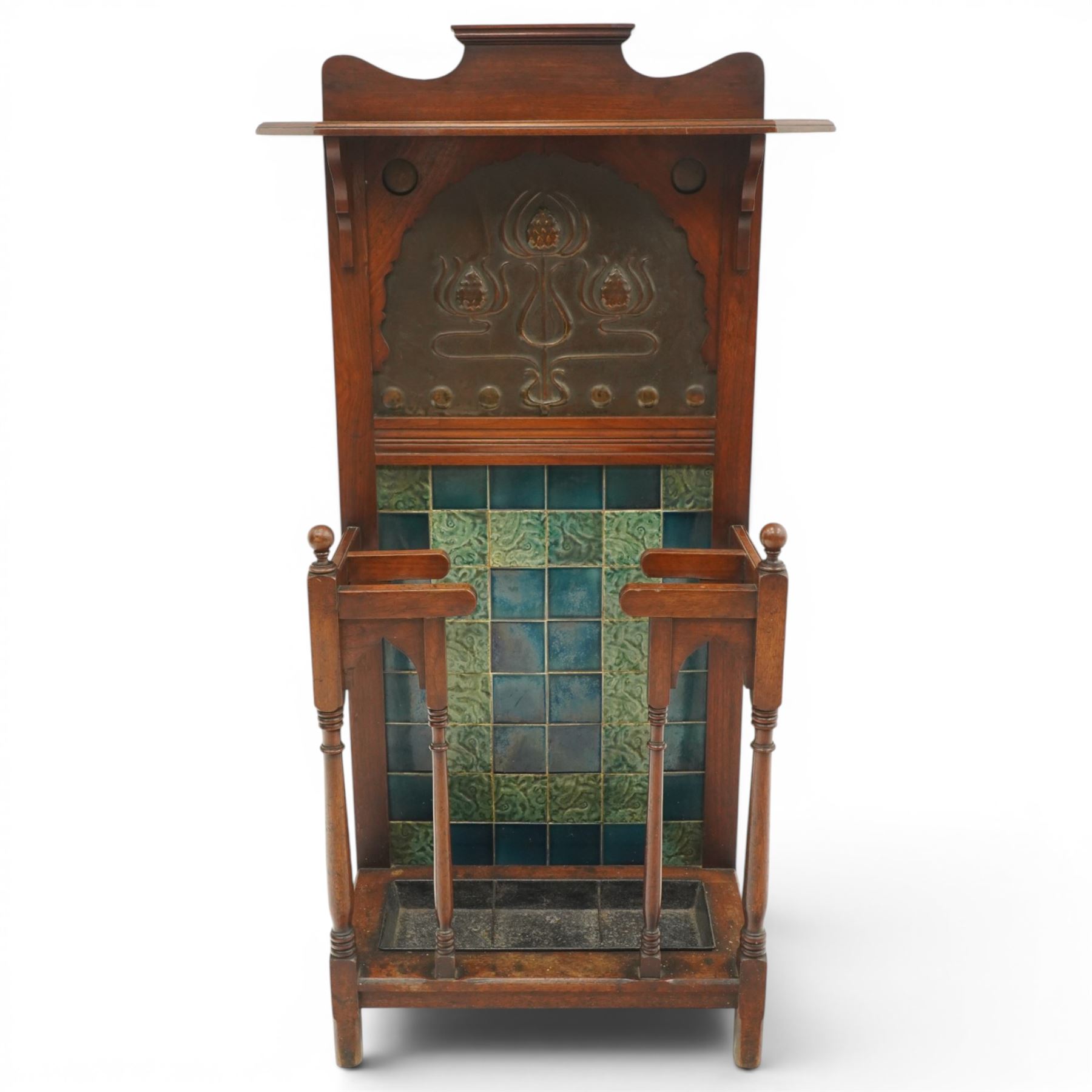 Shapland & Petter of Barnstaple - Arts & Crafts mahogany tile-backed umbrella or stick hall-stand, model no.114, the shaped back with single shelf over an Art Nouveau embossed patinated copper panel and turquoise tiled back, with turned supports and dividers over inset metal drip tray

Notes: A comparable Shapland & Petter stick stand of this model is recorded in Daryl Bennett, Shapland & Petter Ltd of Barnstaple, Museum of Barnstaple and North Devon, p.84, cat. no.5.37, and the model is also listed in Bennett, Liberty’s Furniture 1875-1915, p.298, cat. no.8.6. Examples of this model are known to have been retailed by Liberty & Co.