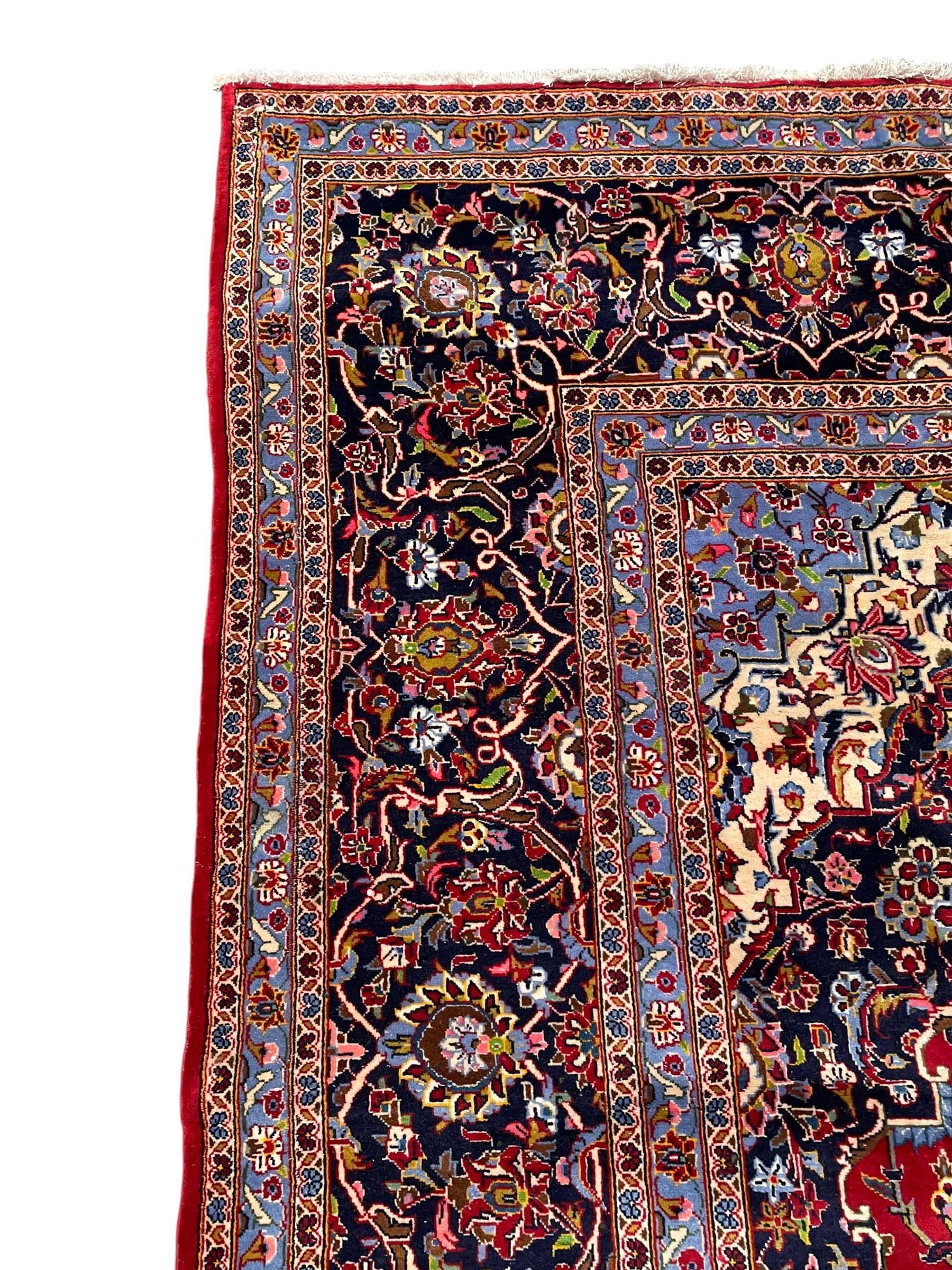 Persian Kashan crimson ground carpet, floral pattern indigo ground central medallion and matching spandrels, the field decorated densely with scrolling foliate patterns and floral motifs, the main border decorated with scrolling branches and palmettes, within floral pattern guard stripes