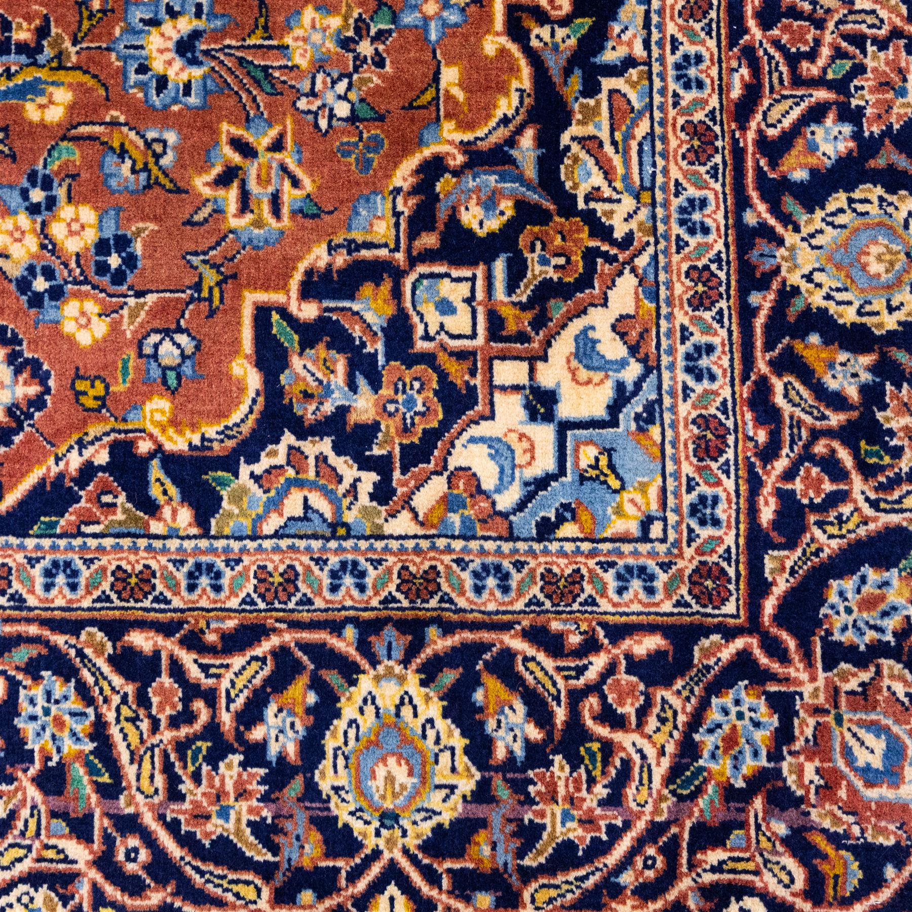 Central Persian Kashan hand-knotted rug,  terracotta field with central shaped ivory and dark blue medallion with palmettes, floral sprays and scrolling vine ornament, with conforming corner spandrels enclosed by a broad navy border of palmettes and flowering vine between multiple guard stripes, with cotton fringe ends 