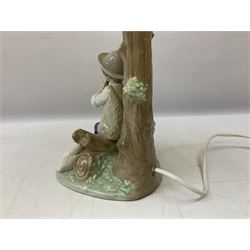 Nao table lamp modelled as a boy playing the flute upon logs with two recumbent sheep at his feet, the stem modelled as a tree trunk, with fabric shade, with printed mark beneath, H46cm