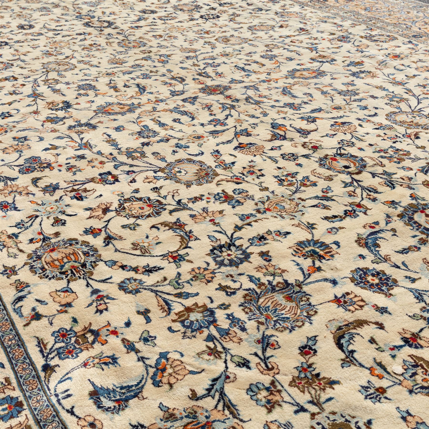 Large Central Persian Kashan hand-knotted carpet, ivory field with an all-over scrolling design of vines, palmettes, and floral sprays, enclosed by a broad camel-ground palmette and vine border, framed by multiple guard stripes in a palette of ivory, beige, blue and polychromed tones, with cotton fringe ends 