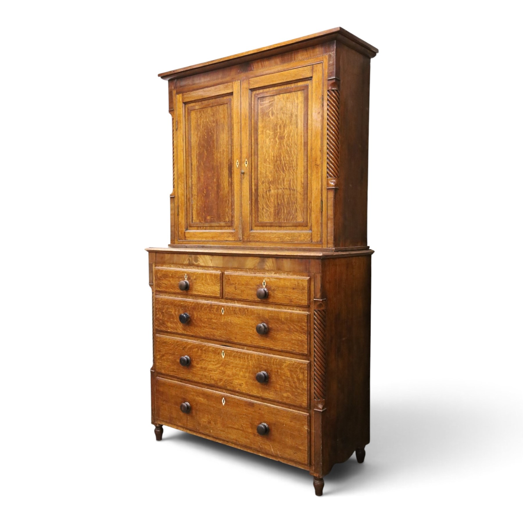Georgian oak and mahogany housekeepers cupboard, the upper section with projecting cornice, crossbanded and inlaid frieze, above panelled doors with bone escutcheons, enclosing three fixed shelves, flanked by barley twist quarter pilasters, the lower section with further crossbanded and inlaid frieze, two short over two long graduating drawers with ebonised handles, raised on turned supports 