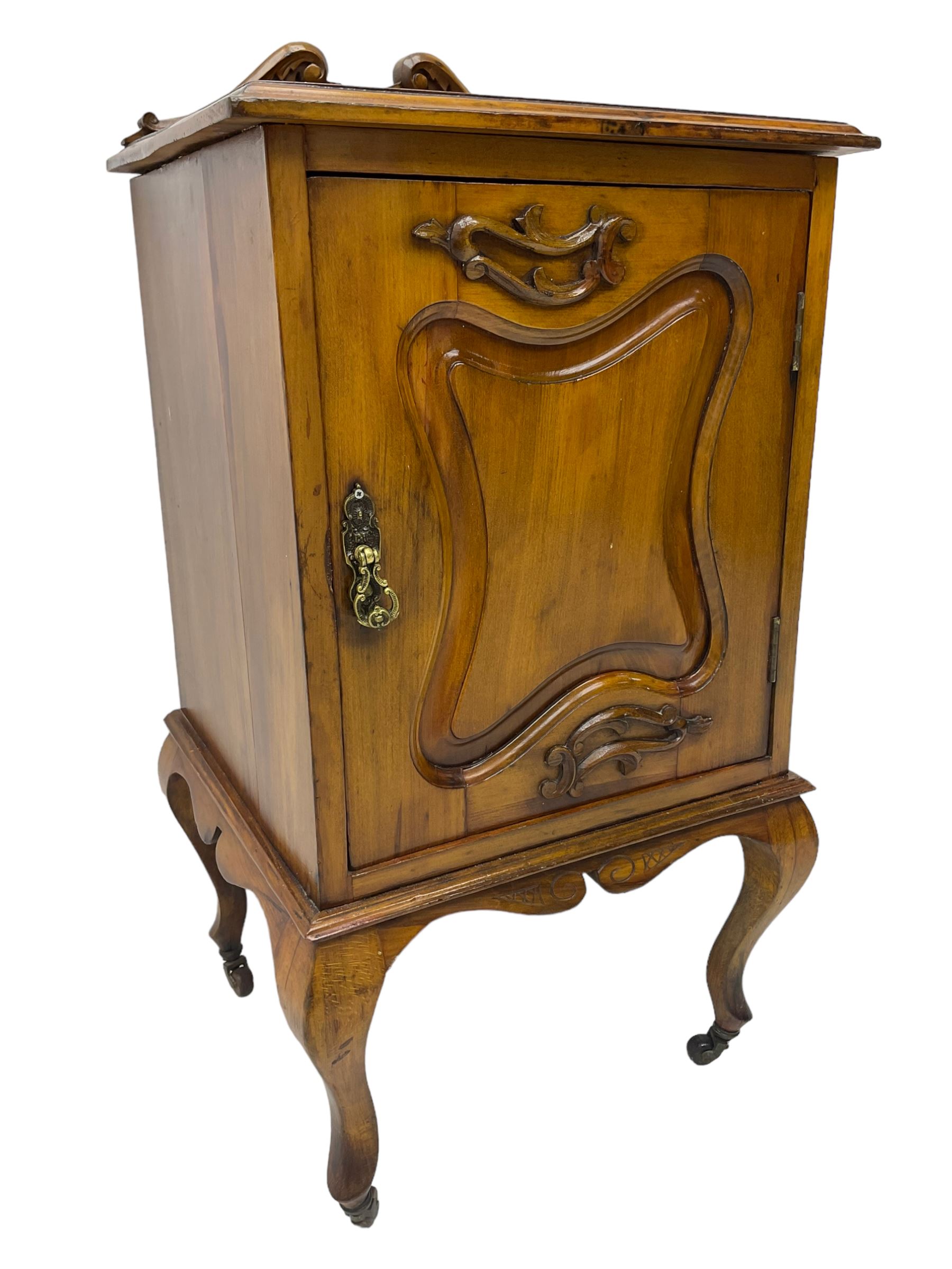Edwardian Art Nouveau walnut bedside cabinet, raised broken swan neck back with carved flower heads, single fielded panelled door enclosing single shelf, raised on cabriole supports with ceramic castors