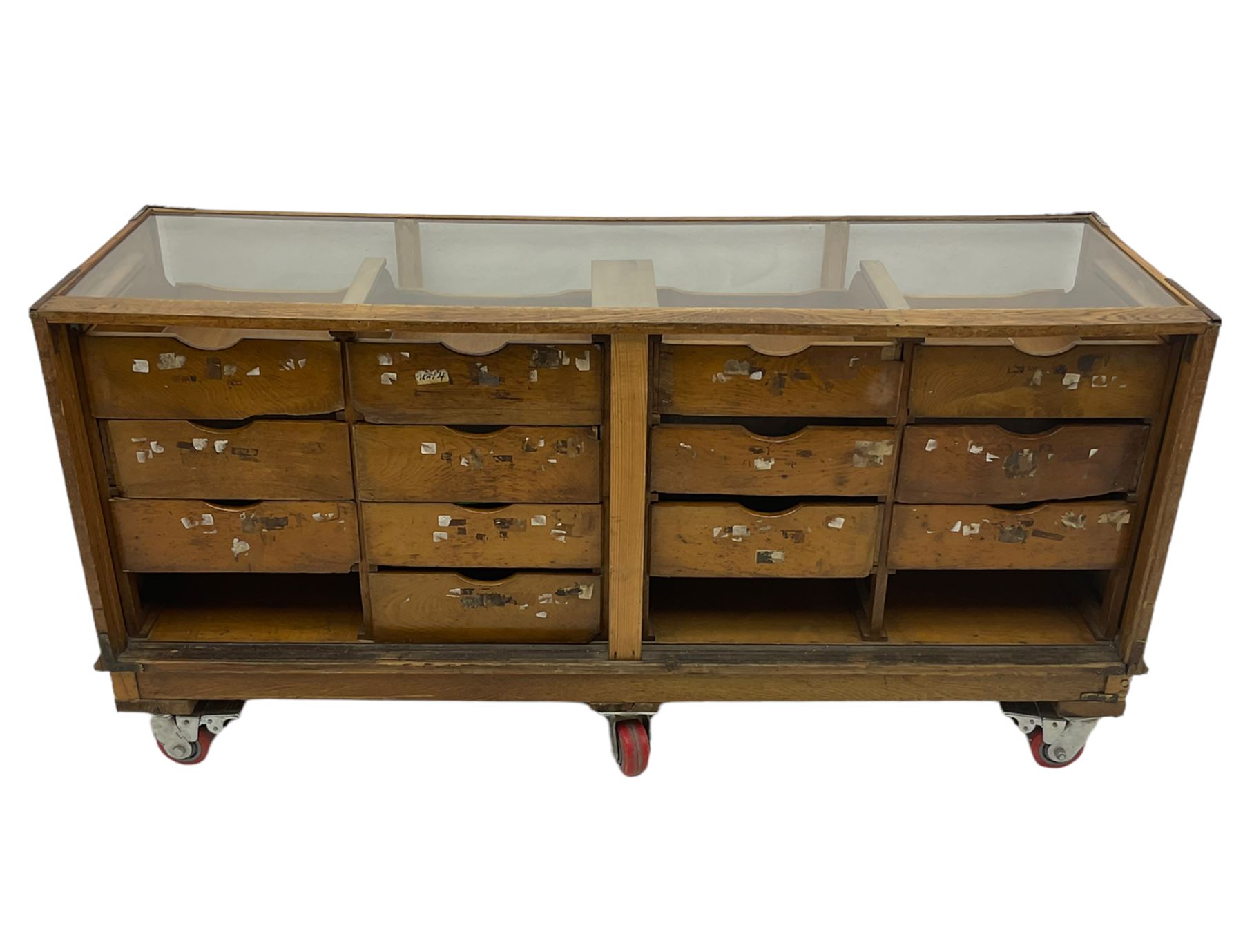 Early 20th century oak framed and glazed haberdashery / shop display cabinet, fitted with sixteen drawers (three missing), on later casters