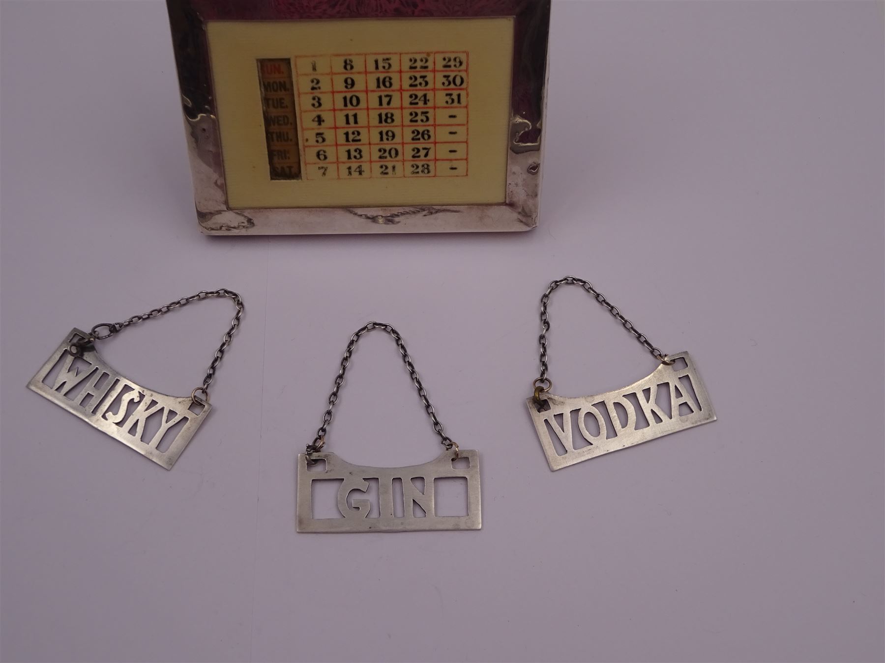 1920s silver mounted perpetual desk calendar, printed in red and black on an ivorine ground, hallmarked Charles S Green & Co Ltd, Birmingham 1925, with wooden easel style support verso, together with a set of three modern silver decanter labels, with cut-out lettering for gin, whisky and vodka, hallmarked London 1982, maker JKM, calendar H8.4cm