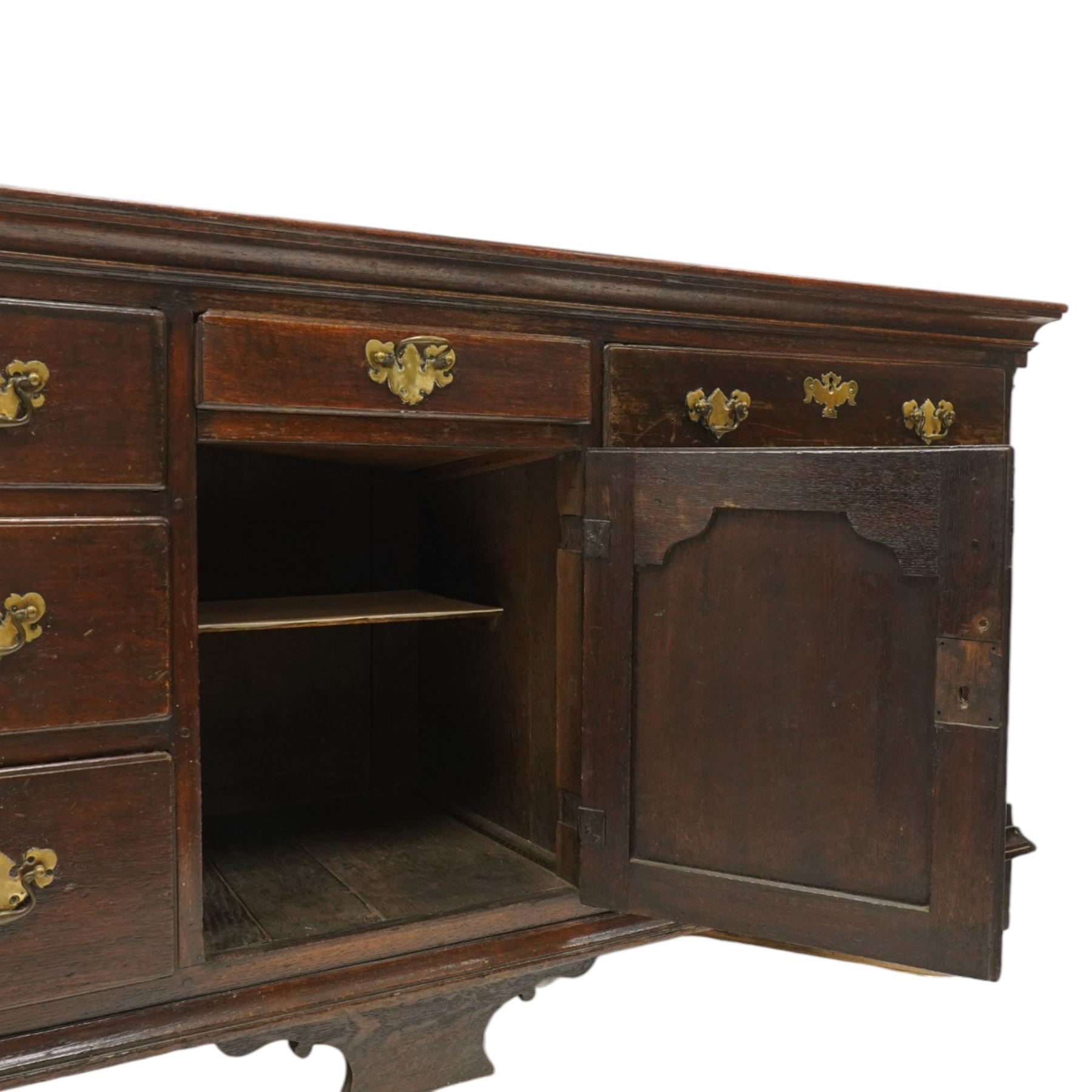 19th century Georgian oak long dresser base, rectangular plank top above a central frieze drawer over a fielded panel cupboard, flanked by three banks with quarter cantered pilasters, brass swan neck handles and escutcheons, raised on shaped bracket feet