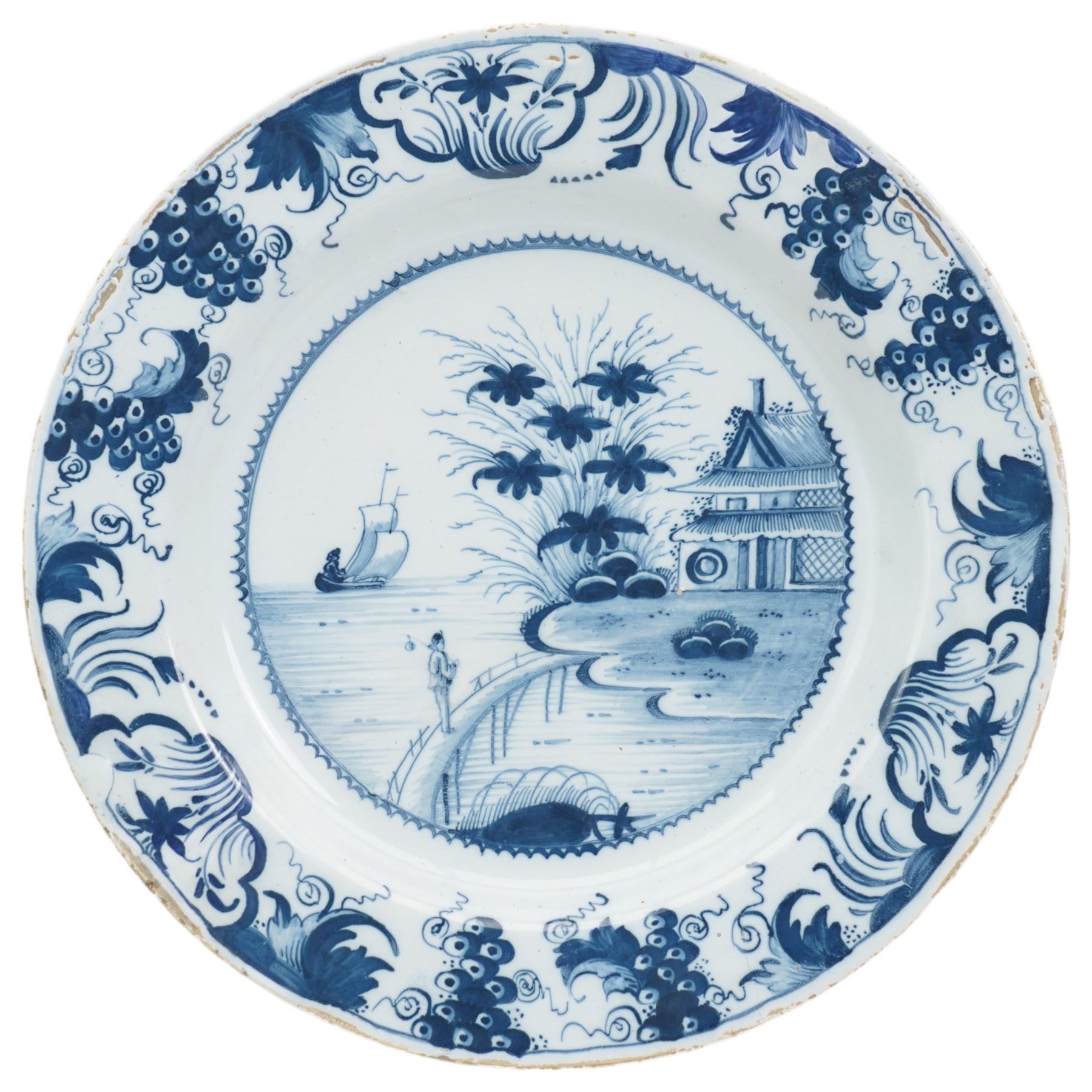 18th century English Delft charger, painted in blue with a Chinese scene with a figure in a boat, another figure crossing a bridge towards a tree and house, within a border of grapes and foliate scrolls, a similar pattern was produced by Delftfield, Glasgow, c.1760, D41cm