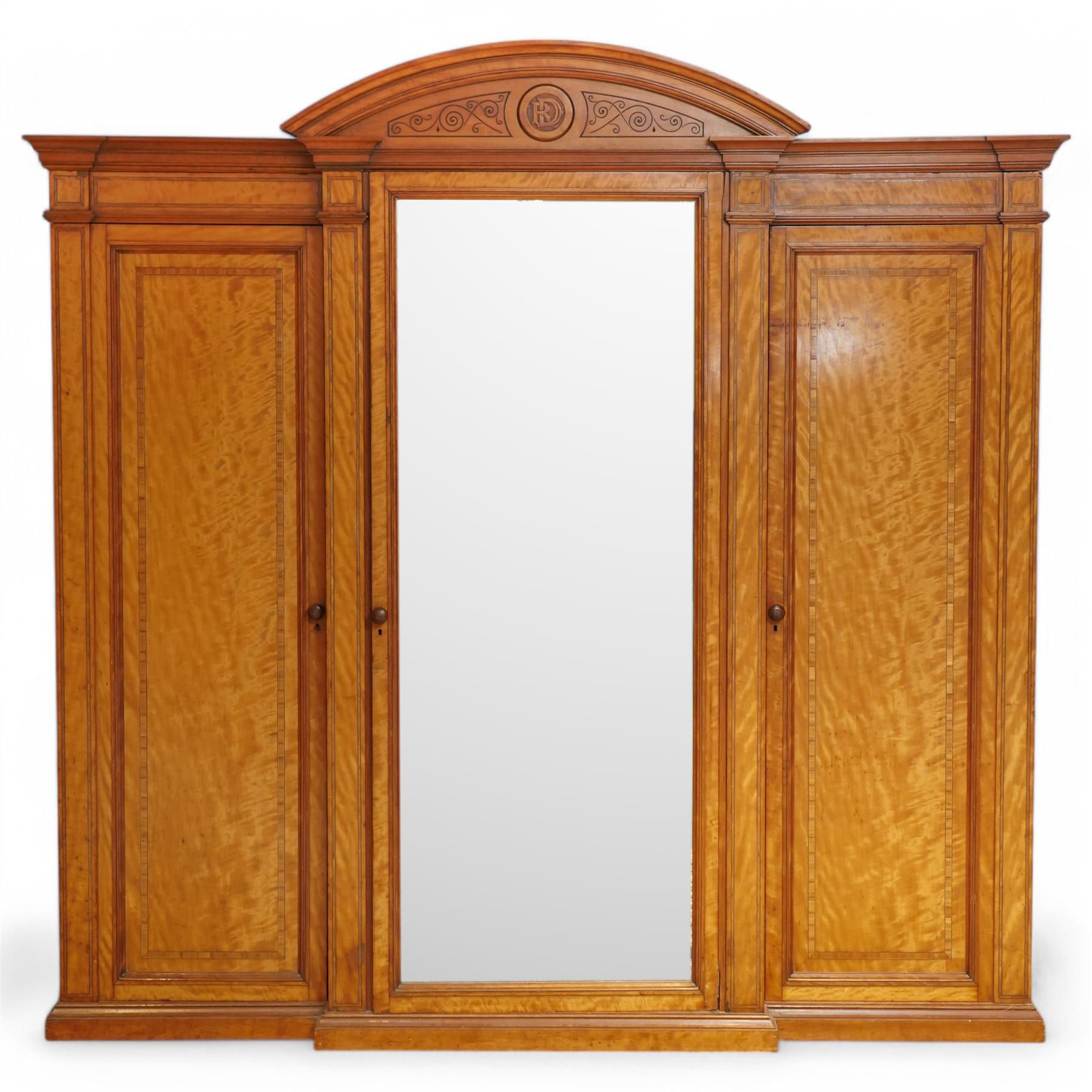 Victorian neoclassical style satinwood triple-section wardrobe, the central mirror section and arched pediment with carved monogram flanked by two panelled crossbanded doors, revealing two drawers and grooves for shelves, the hanging cupboards each with a single drawer, the whole supported on a plinth base