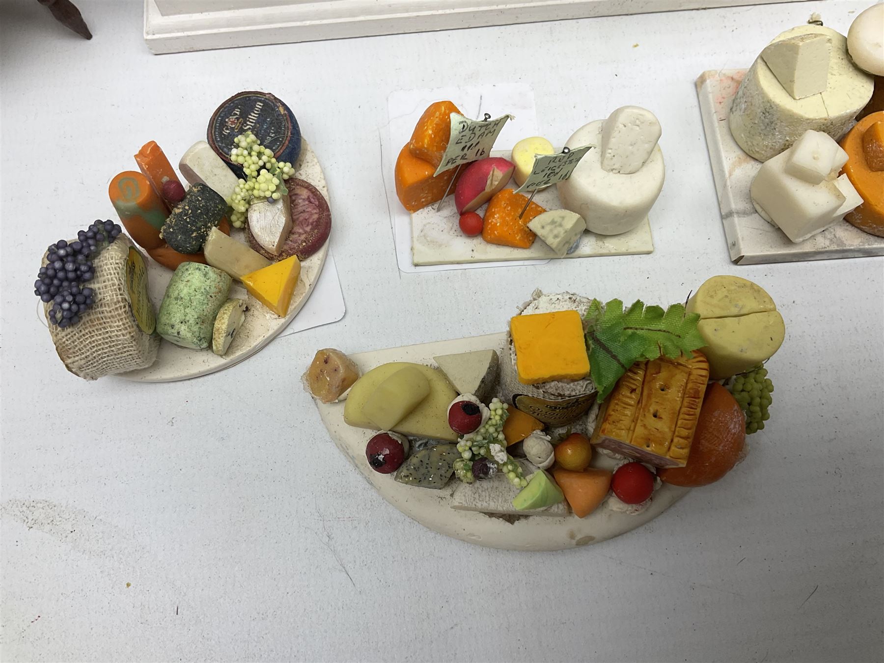 Collection of miniature dolls house grocery shop furniture and produce, comprising glazed corner counter with brass style cash register, shelved preserves unit, pies, cheese board and trays of meats, pair of white cheese counters, biscuit display with six tins, glazed preserves cabinet, pair of shop chairs and four further cheese boards complete with grapes and label flags