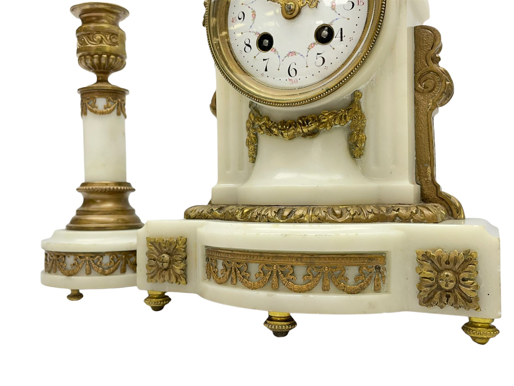 An early 20th century white marble mantle clock in a break front case with an arched top and finial, gilded scroll work to the sides and pierced foliate panels to the front, plinth raised on five bun feet, enamel dial with garland decoration, Arabic numerals, minute markers and Louis XV gilt hands, within a convex glazed bezel, eight-day Parisian striking movement striking the hours and half hours on a bell, with two conforming single light candlesticks. With Pendulum.

