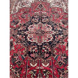 Persian Heriz red ground rug, central scrolled medallion on dark ground decorated with floral motifs, the surrounding field decorated with stylised plant motifs and smaller geometric symbols, guarded border with repeating pattern punctuated with rosettes and flower heads 