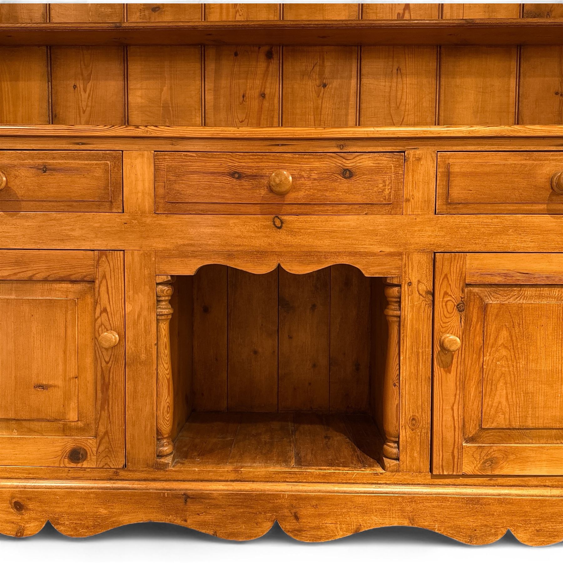 Late 20th century pine dresser, the plate rack with two fixed shelves and shaped apron over panelled back, base fitted with three drawers over two cupboard doors and central open niche flanked by turned uprights, on plinth base with shaped apron