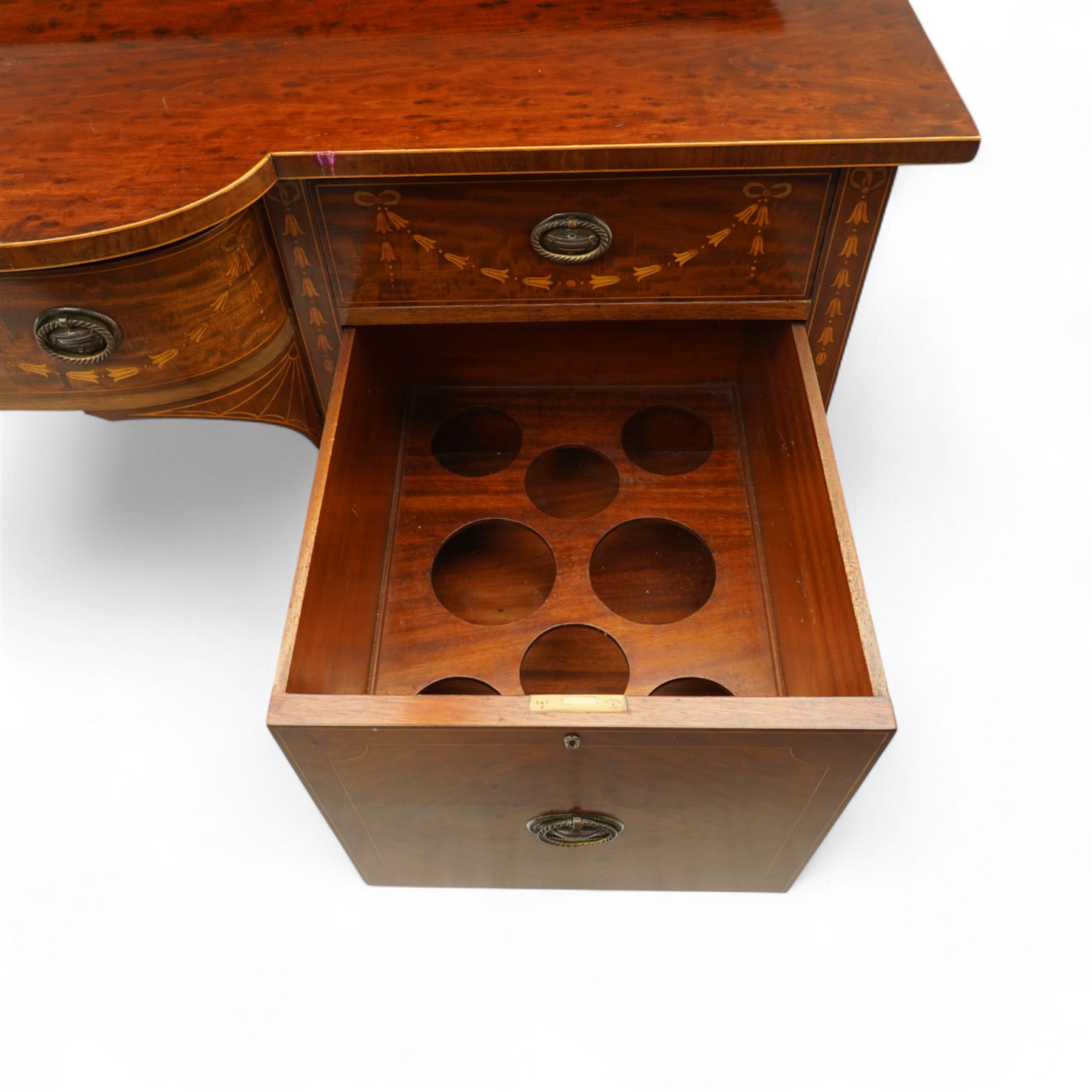 Edwardian Sheraton Revival inlaid plum pudding mahogany serpentine sideboard, rectangular top with raised gallery above bowed frieze drawer finely inlaid with central neoclassical urn motif, flanked by two drawers with swags and ribbon bows, above a cellarette drawer and cupboard, on square tapered supports with spade feet 