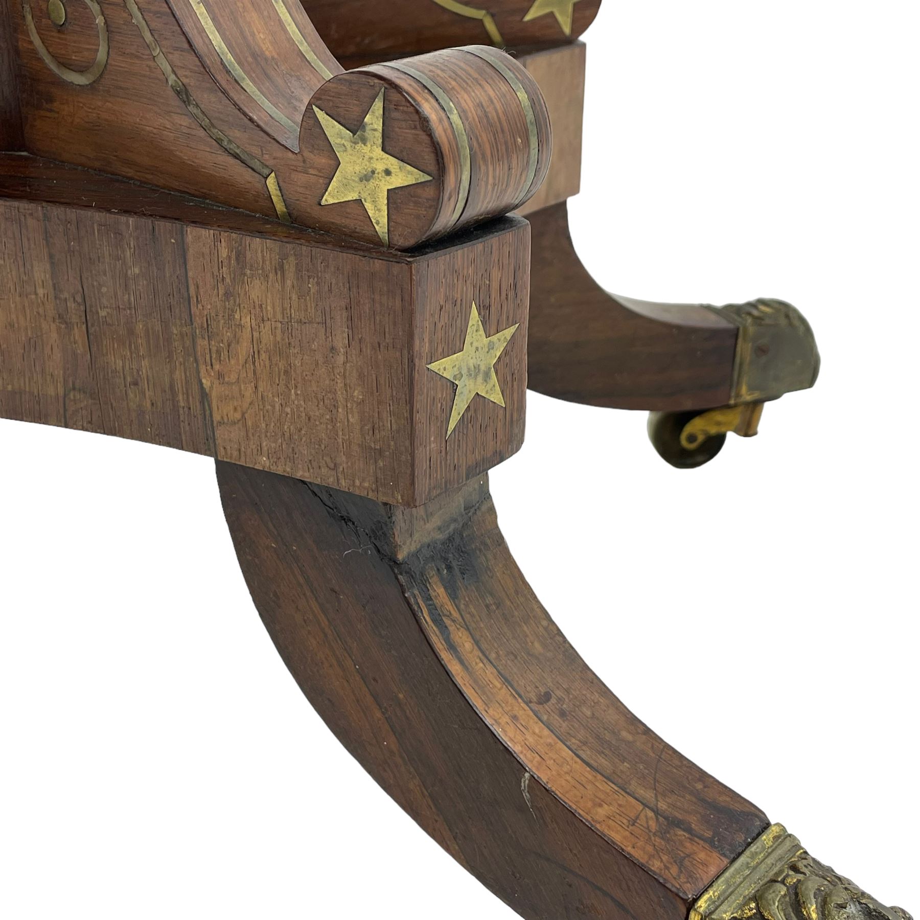 Regency rosewood and brass inlaid card table, the rectangular fold-over and swivelling top with rounded corners with a satinwood band, opening to reveal a blue baize lined interior playing surface, the frieze with a raised panel with satinwood stringing and a lower beaded brass edge, the concave and canted column with a shaped platform base applied with scrolls with brass star inlays and stringing, quadruple splayed supports terminating in foliate cast cups and castors