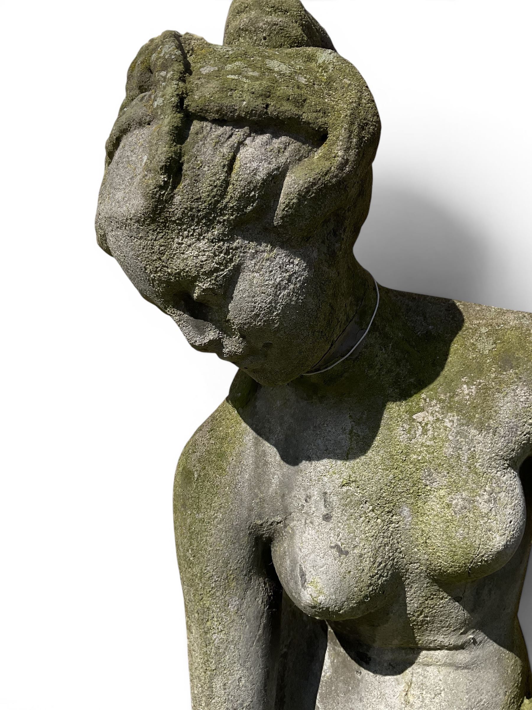 Cast stone garden figure of a classically draped nude female inspired by Aphrodite statues, with stylised up-do arranging her chitin on a circular base, the rectangular pedestal with egg-and-dart moulding over a floral wreath, in weathered finish