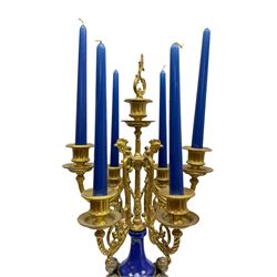 A 20th century continental gilt metal Lyre mantle clock with a pair of matching six light candelabra, spring driven movement housed in a gilt drum case on a brass and cobalt blue base with paw feet, two bronzed metal figures of mythological cherubs with animal legs and cloven hooves supporting a festooned garland swag, eight-day twin barrel striking movement with a floating lever balance escapement, striking the hours and half-hours on two bells, white enamel dial with roman numerals, minute track and pierced steel hands, dial inscribed “Imperial”. 