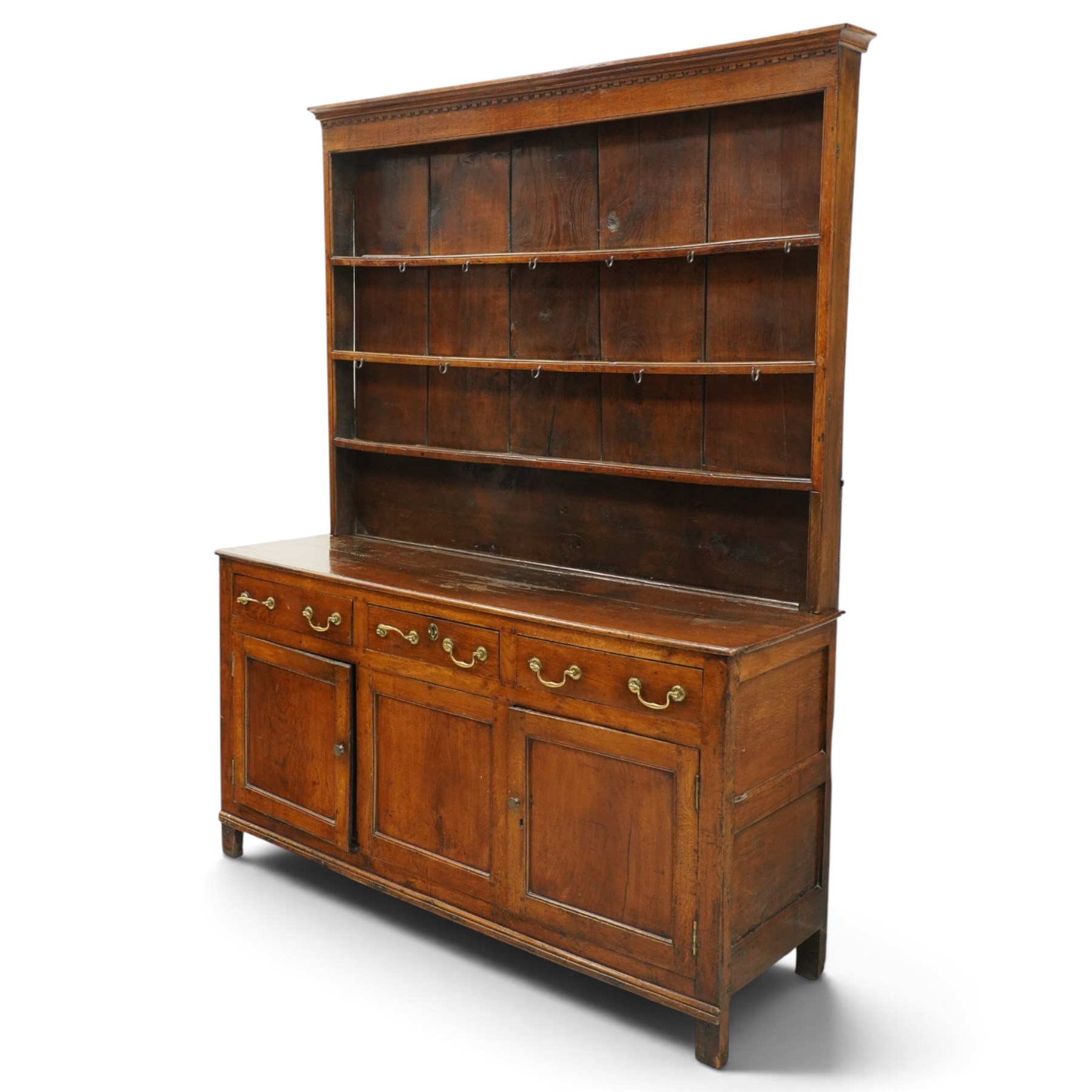 18th century oak dresser with rack, the three-tier plate rack with dentil-carved cornice, the plate shelves with moulded edge and wrought metal hooks, the base fitted with three drawers with brass swan-neck handles over three panelled cupboards, lower moulded edge on stile supports