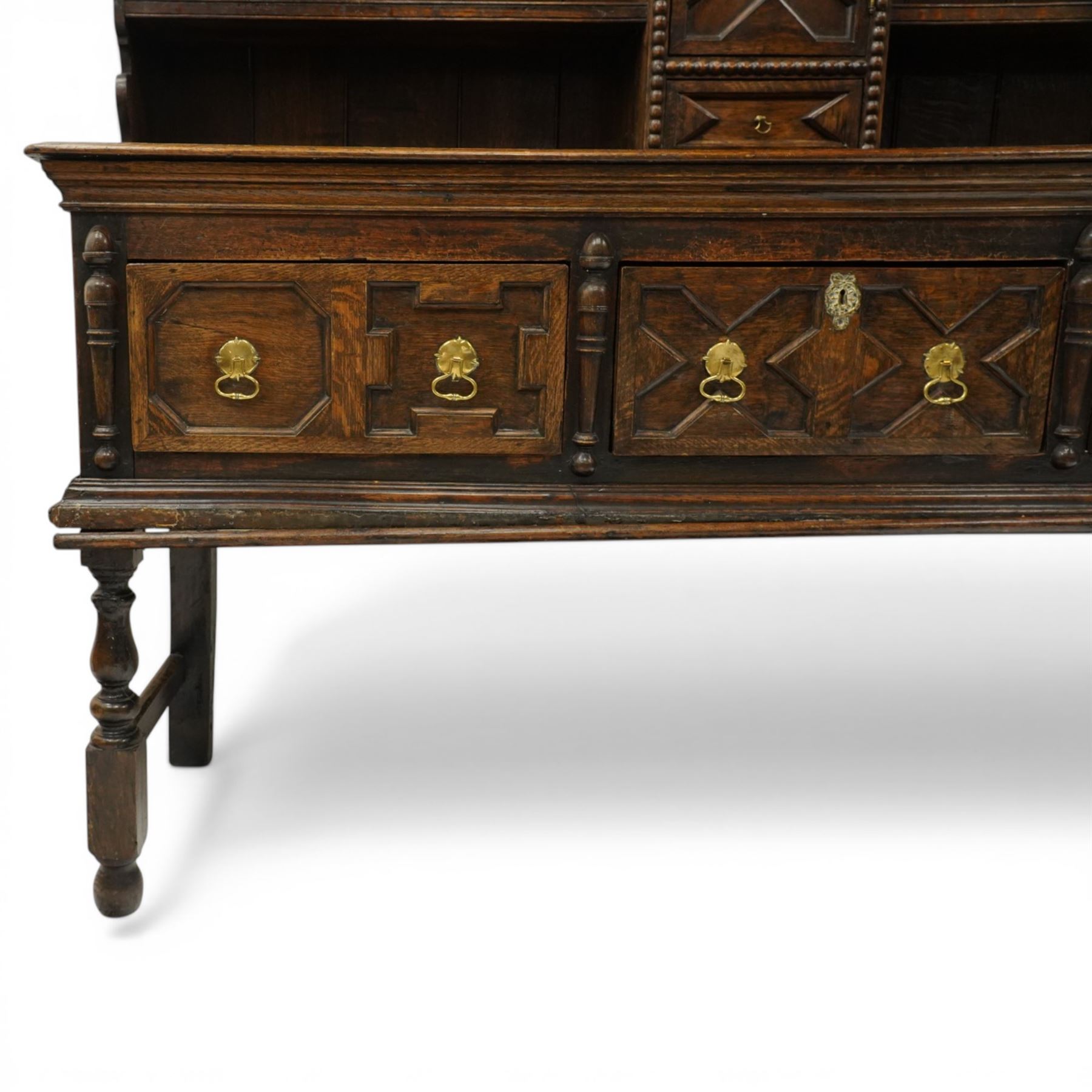 19th century Jacobean Revival oak dresser and rack, the upper section with open shelves and central cupboard, flanked by bobbin moulded uprights, the base fitted with three carved drawers with brass ring handles, raised on turned supports united by stretchers