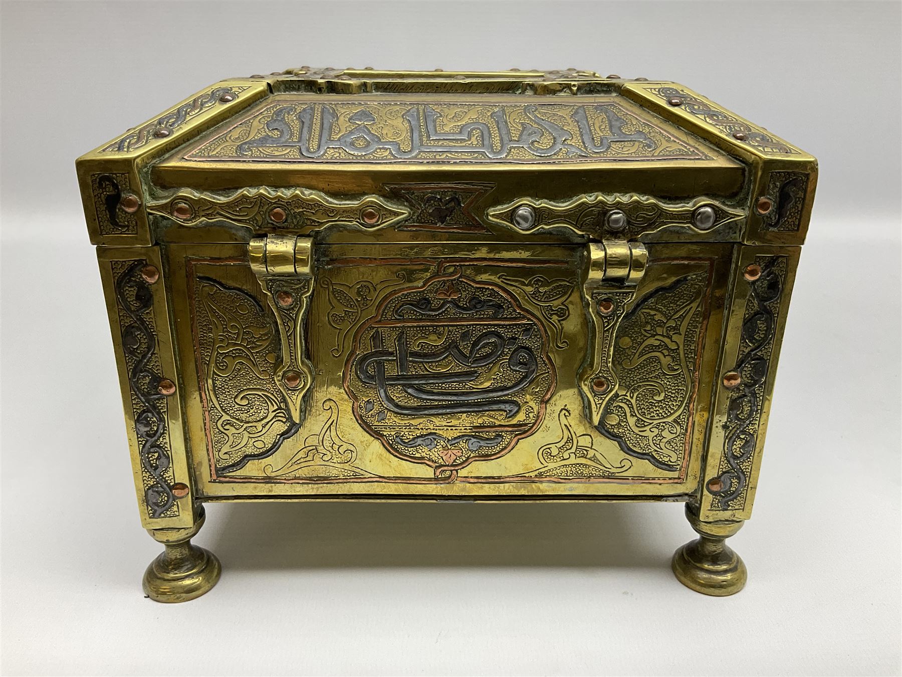 Late 19th century Damascus Cairoware Quran box, of square form with canted hinged cover, upon four circular pad feet, the brass exterior inlaid with silver and copper script and decorative detail, opening to reveal a marquetry lined interior, H15.5cm W19cm D19cm