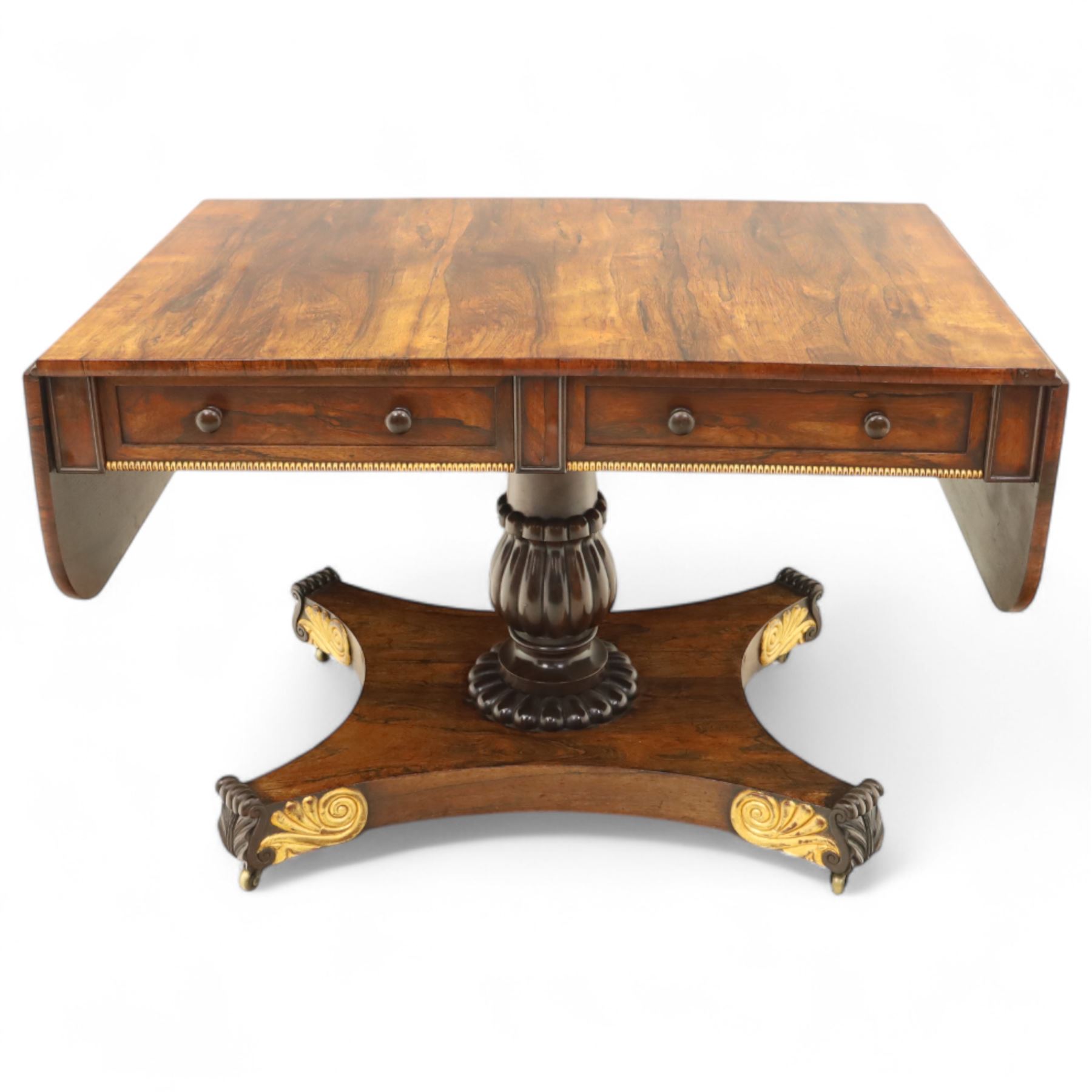 Regency rosewood sofa table, rectangular top with variegated rosewood veneers, hinged D-shaped leaves, over a set of dummy drawers to the back and full-size drawers to the front, with turned knob handles and gilt beaded moulding on frieze above elegant turned, tapering, and fluted, pedestal with a well-carved collar, supported on a large, tripartite concave platform base terminating in richly carved lion paw feet with gilded acanthus leaf carving, on concealed brass castors, circa 1815, length extended 161cm 
