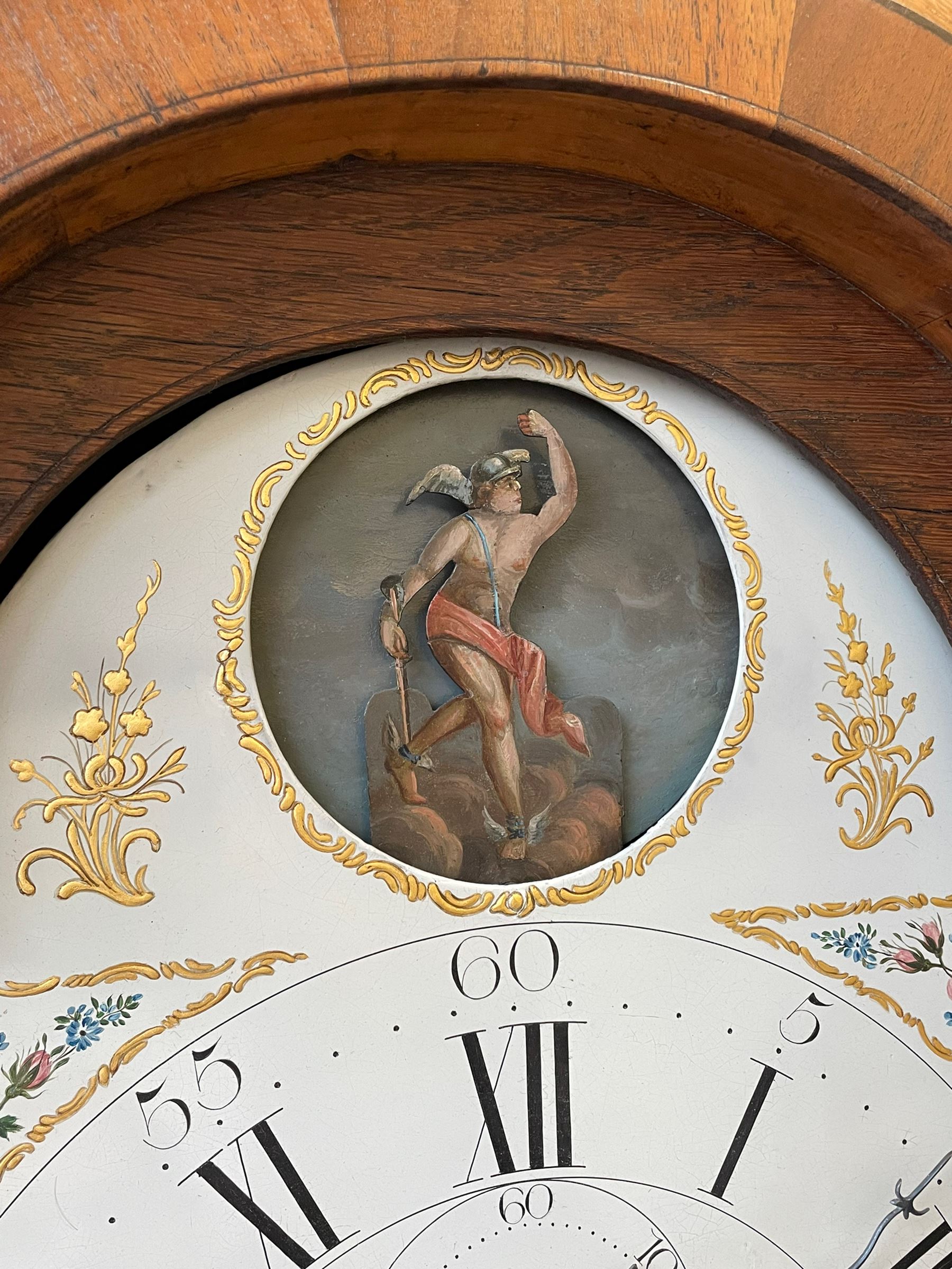 William Gostling of Diss  (Norfolk)-  Late 18th century 8-day oak longcase clock c1780-90, break arch top with scalloped cresting and three brass finials, glazed break arch hood door flanked by turned pilasters with brass capitals, trunk with a long trunk door and rounded top on a square plinth with an applied and shaped skirting, early painted Wilson of Birmingham dial with Roman numerals and five minute Arabic's, matching steel hands and second pointer, arched square ended calendar aperture with date disc behind, floral spandrels within gesso borders and a painted rocking automaton representing Mercury to the break arch, dial pinned via a false plate to a weight driven rack striking movement with a recoil anchor escapement, striking the hours on a cast bell. With pendulum weights and key. 
William Gostling (II) is recorded as working in Diss from 1774 to 1797, succeeded by widow Anne and son George . The dial of this clock is illustrated and discussed in detail in M.F. Tennants book 