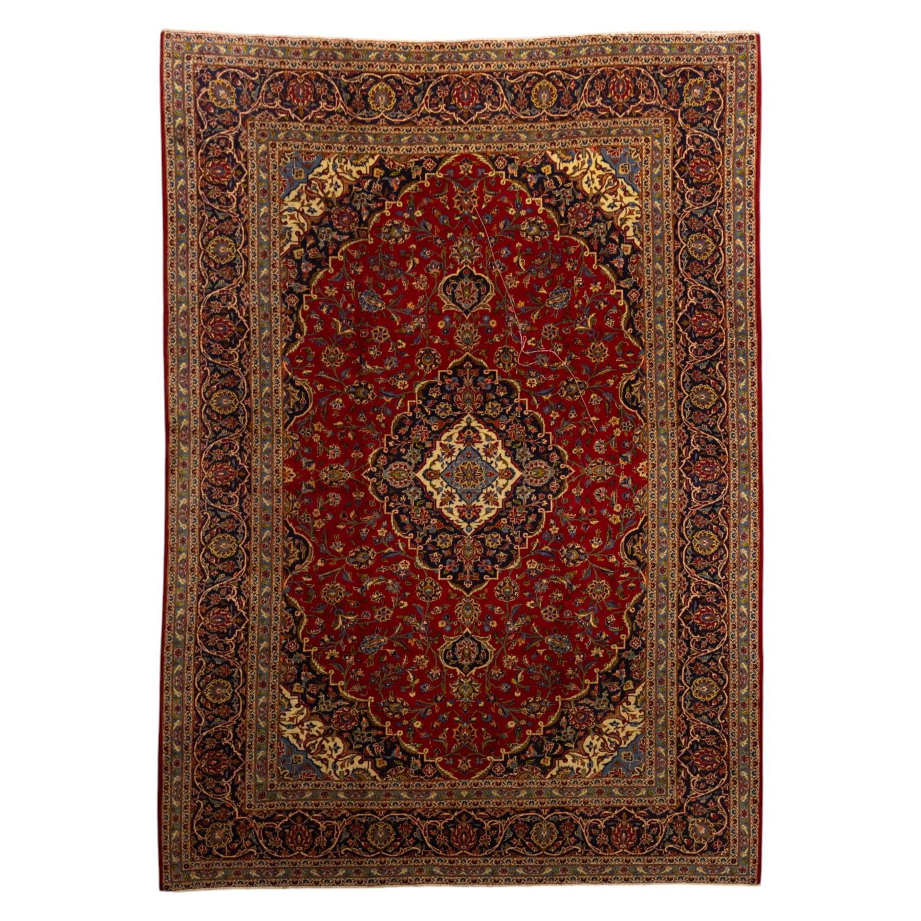 Central Persian Kashan hand-knotted carpet, crimson ground, with a central shaped palmette medallion with pendants, surrounded by scrolling foliate and floral motifs incorporating rosettes, palmettes, and vine work, enclosed by a dark navy with repeating border with repeating palmettes and meandering vines, flanked by floral guard stripes, with cotton fringes 