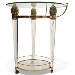 Hollywood Regency style gilt metal and glass serving trolley, circular top in a polished brass surround with three pineapple finials, supported on clear column legs, lower glass shelf enclosed by a brass gallery, terminating in castors