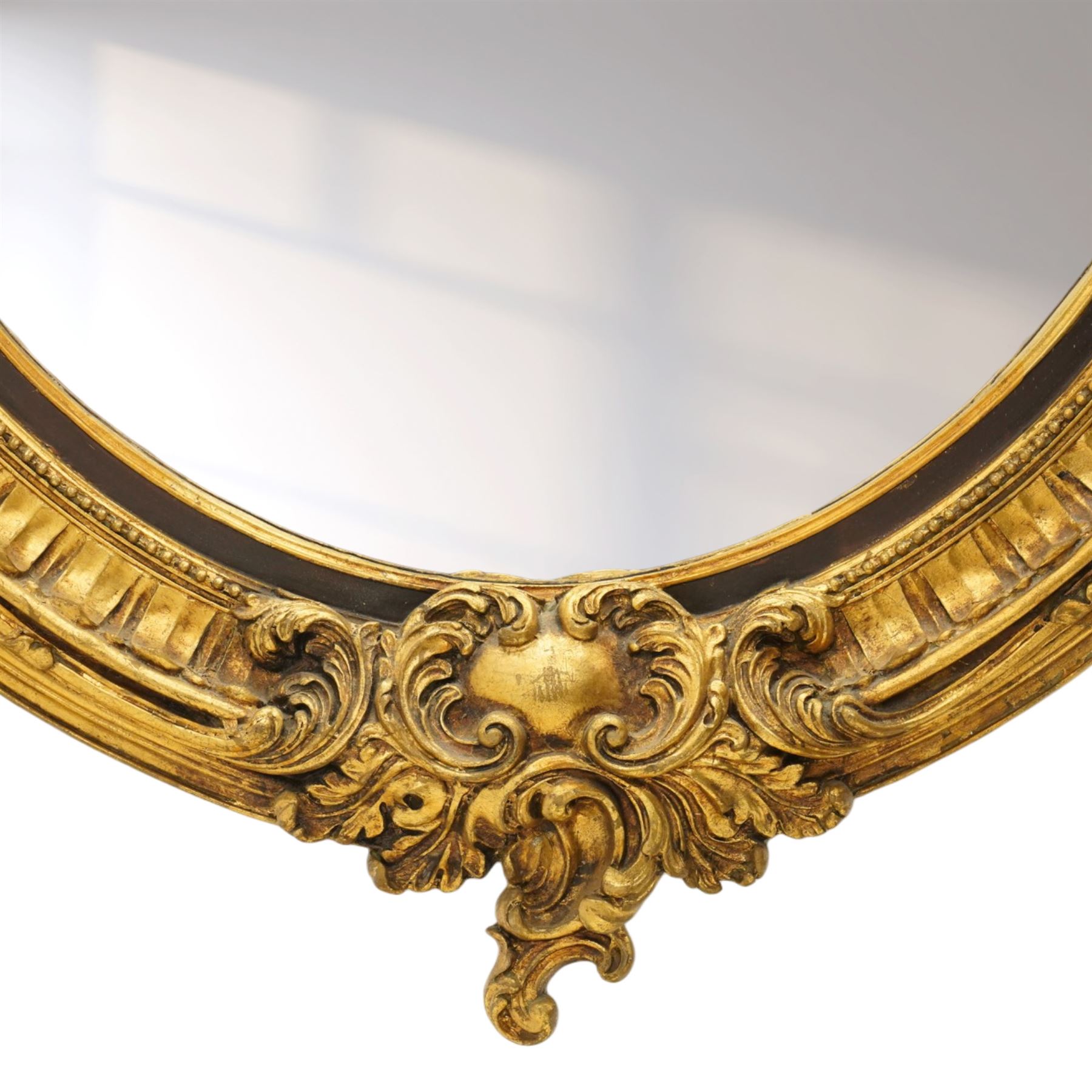 Pair of Rococo Revival gilt framed oval wall mirrors, the exuberant pediment formed of scrolling acanthus leaves and rocaille ornament, the oval bevelled plate set within with ebonised slip with outer repeating lappet decoration, over a rocaille cartouche terminal