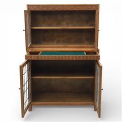 Early 20th-century Arts and Crafts glazed oak bookcase, upper section fitted with a pair of leaded glazed doors enclosing a fixed shelf above a projecting frieze drawer interior with baize-lined writing surface flanked by two compartments, carved with interlaced decoration, lower section fitted with a further pair of leaded glazed doors enclosing a single shelf, raised on short stile feet 